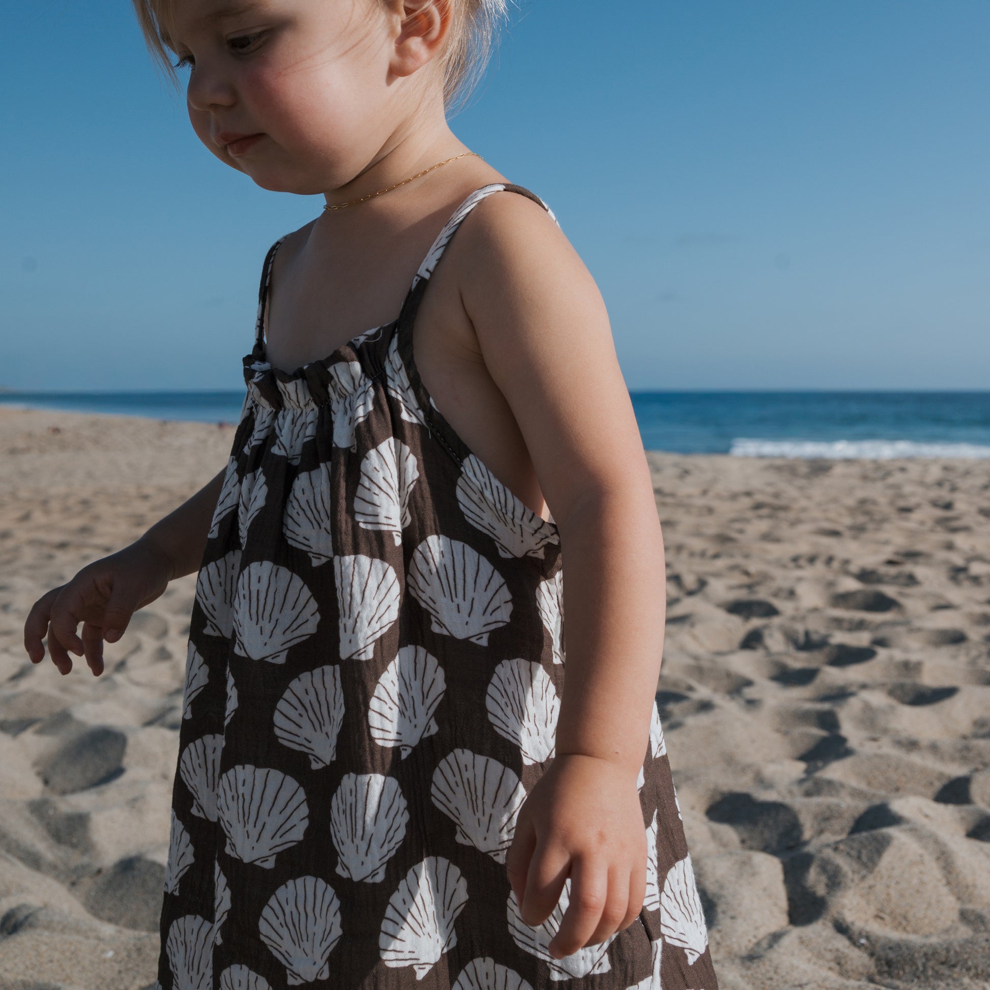 A young child wears the forever french baby Child Muslin Sleeveless Dress | Seashells, featuring white seashell patterns, while standing on a sandy beach with the blue sky and ocean in the background.