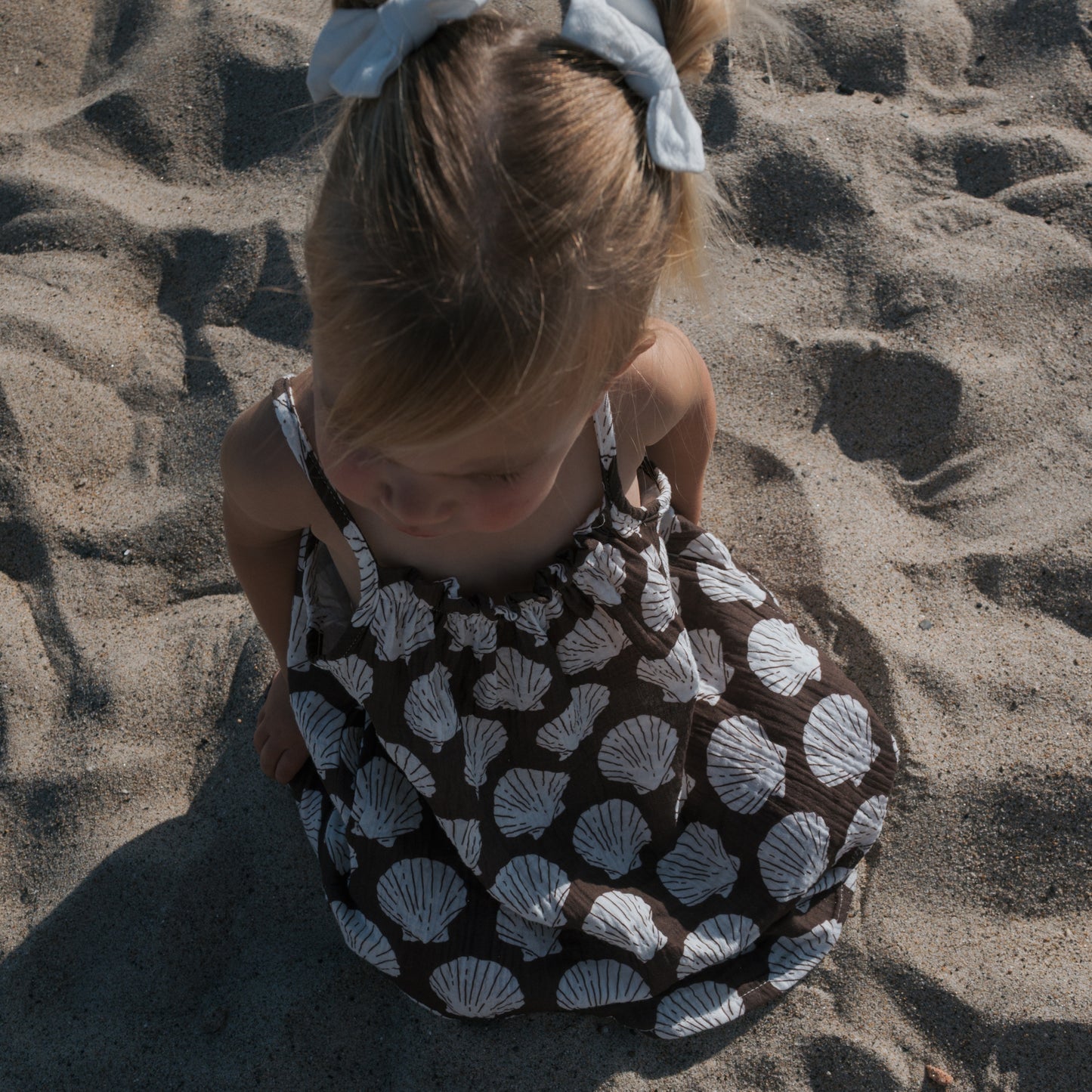 A young girl with a white bow sits on sandy ground, wearing the forever french baby Child Muslin Sleeveless Dress | Seashells, as sunlight casts gentle shadows around her.