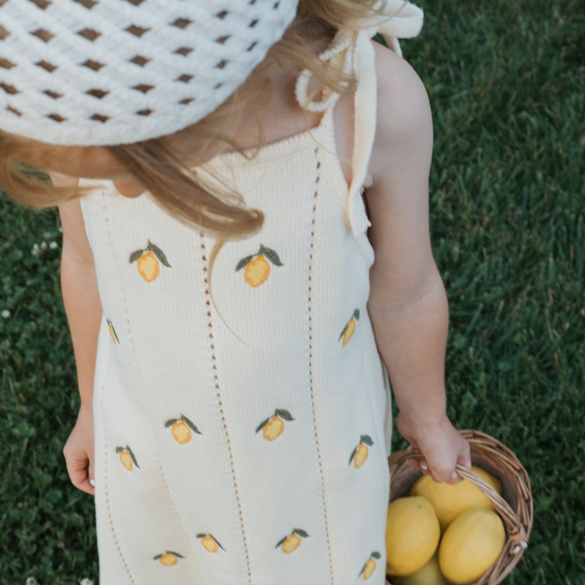 A young child wears the forever french baby Knit Dress | Lemon—100% cotton cream with yellow lemon patterns—along with a white crocheted hat, holding a basket of lemons and standing on green grass, seen from above.