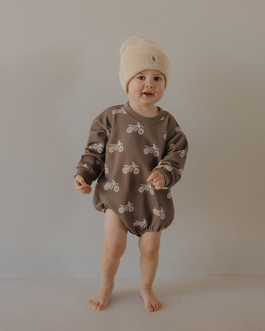 A toddler smiles indoors against a neutral wall, wearing the forever french baby Sweatshirt Romper | Dirt Bike and a beige knit beanie, with hands slightly raised.