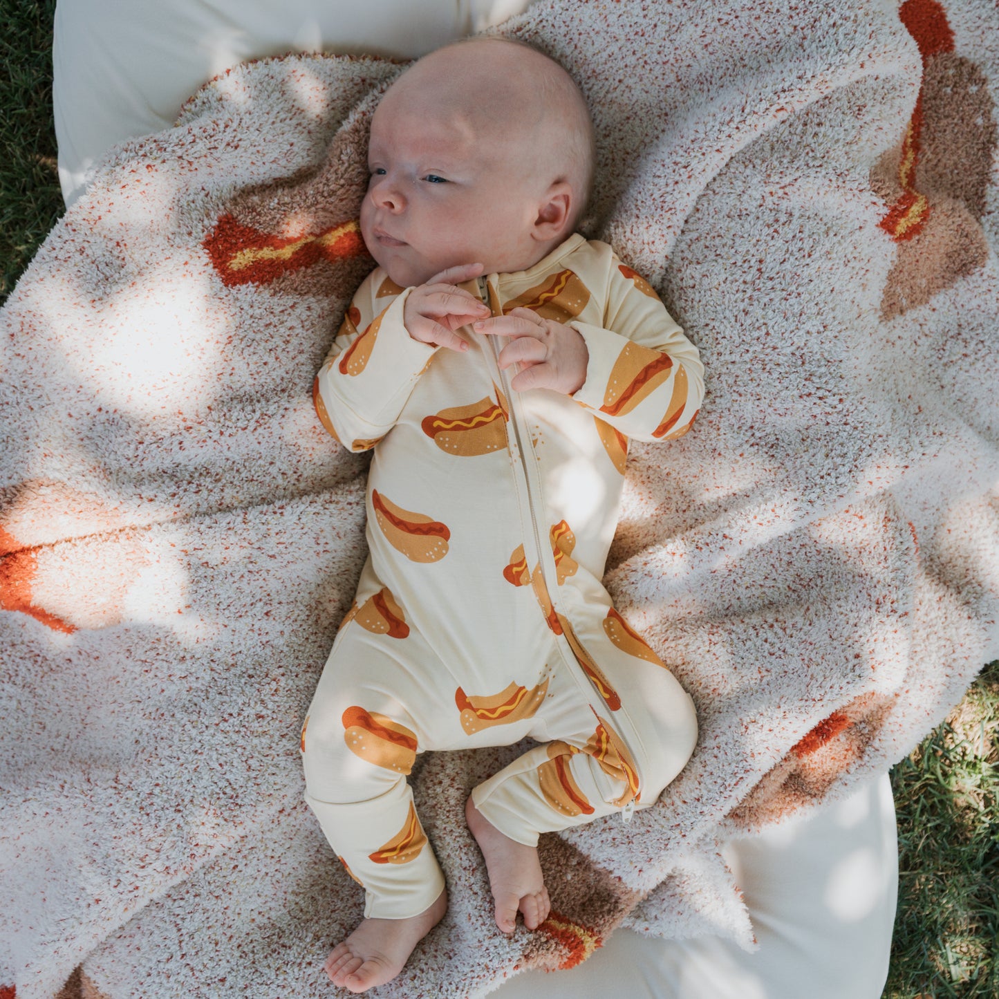 A baby in a hot dog-patterned onesie relaxes on the forever french baby Plush Blanket | Hot Dogs, an Oeko-Tex certified blanket, outdoors and gazes off to the side with hands clasped near their chest.