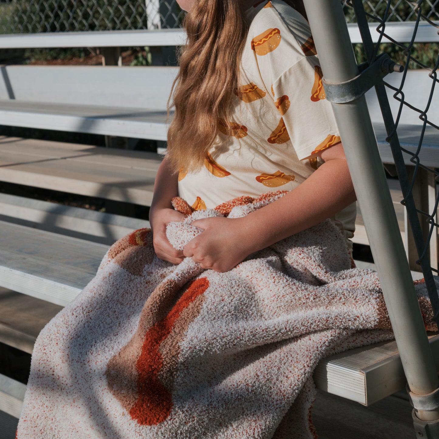 A child with long hair sits on metal bleachers, wearing the forever french baby Girl's Bamboo Short Set | Hot Dogs and holding an orange and beige textured blanket on their lap; a chain-link fence appears behind them.