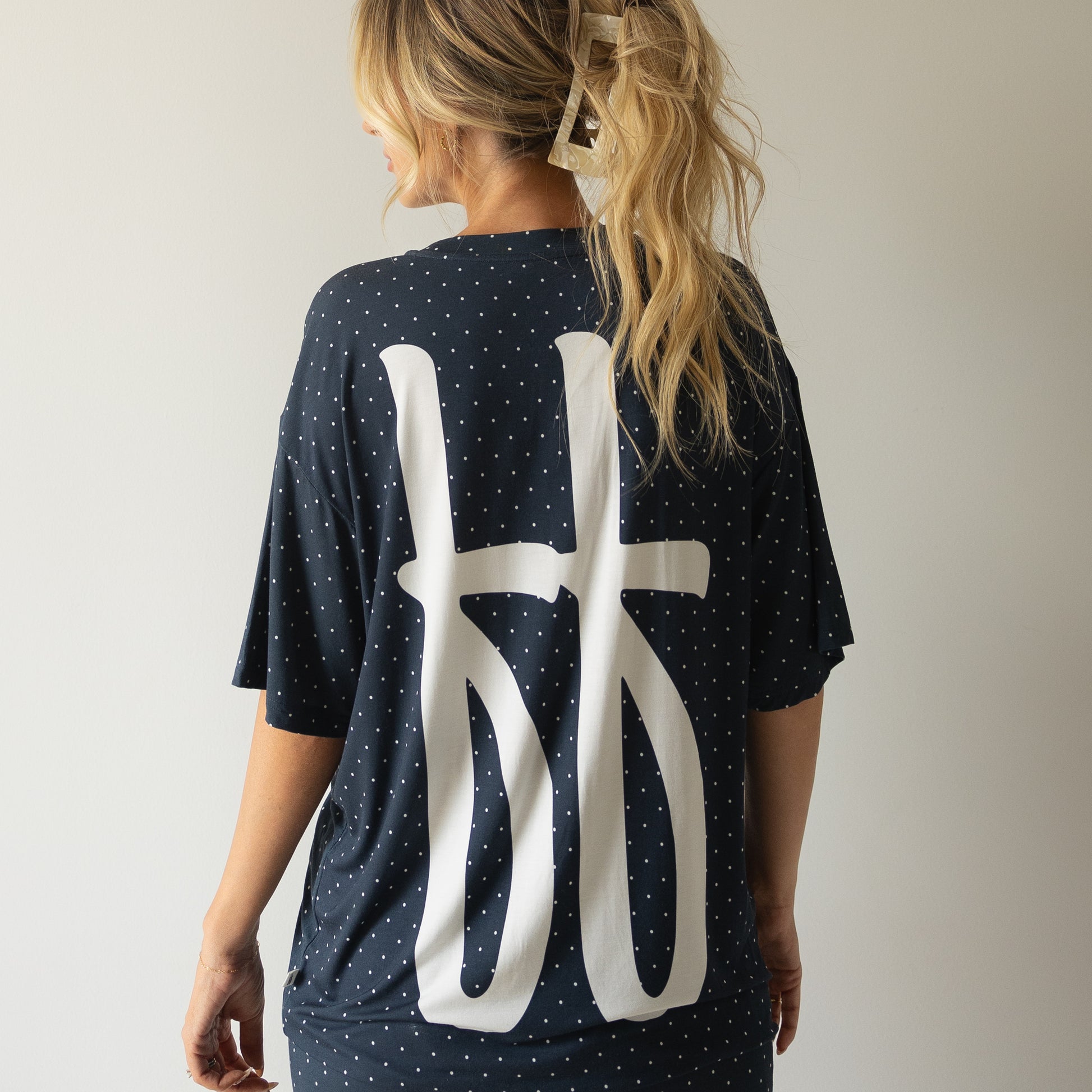 A woman with wavy blonde hair in a clip wears the forever french baby Short Sleeve Women's Bamboo Lounge Set | Emery in navy blue with white polka dots and abstract symbols, standing against a light-colored wall.