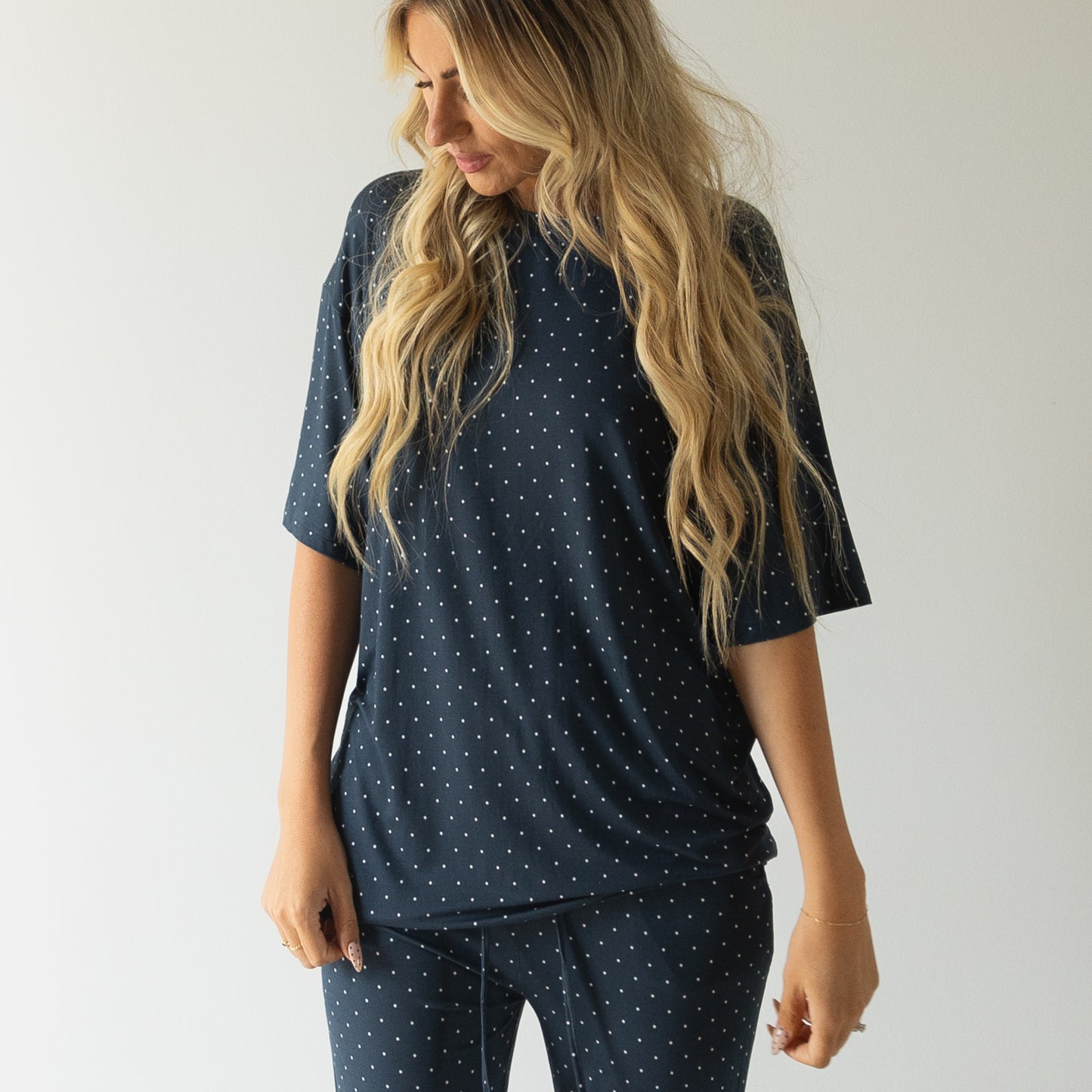 A woman with long, wavy blonde hair wears the forever french baby Short Sleeve Women's Bamboo Lounge Set | Emery in dark blue polka dots, standing against a plain white background and looking down to the side.