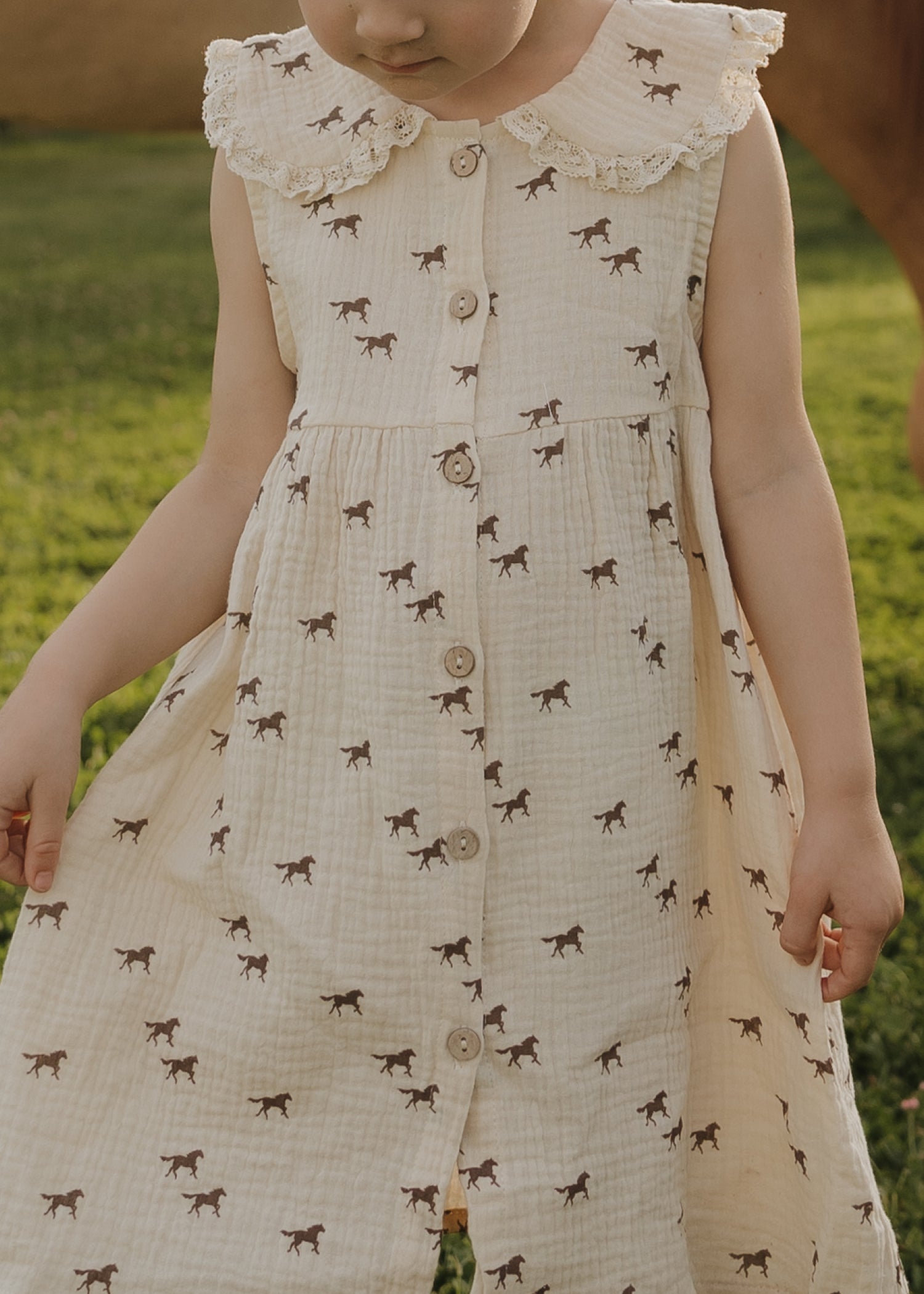 A child in the forever french baby Child Muslin Dress | Wild Horses stands on green grass, looking down with arms slightly out, wearing a sleeveless cream dress with lace collar and brown horse prints.