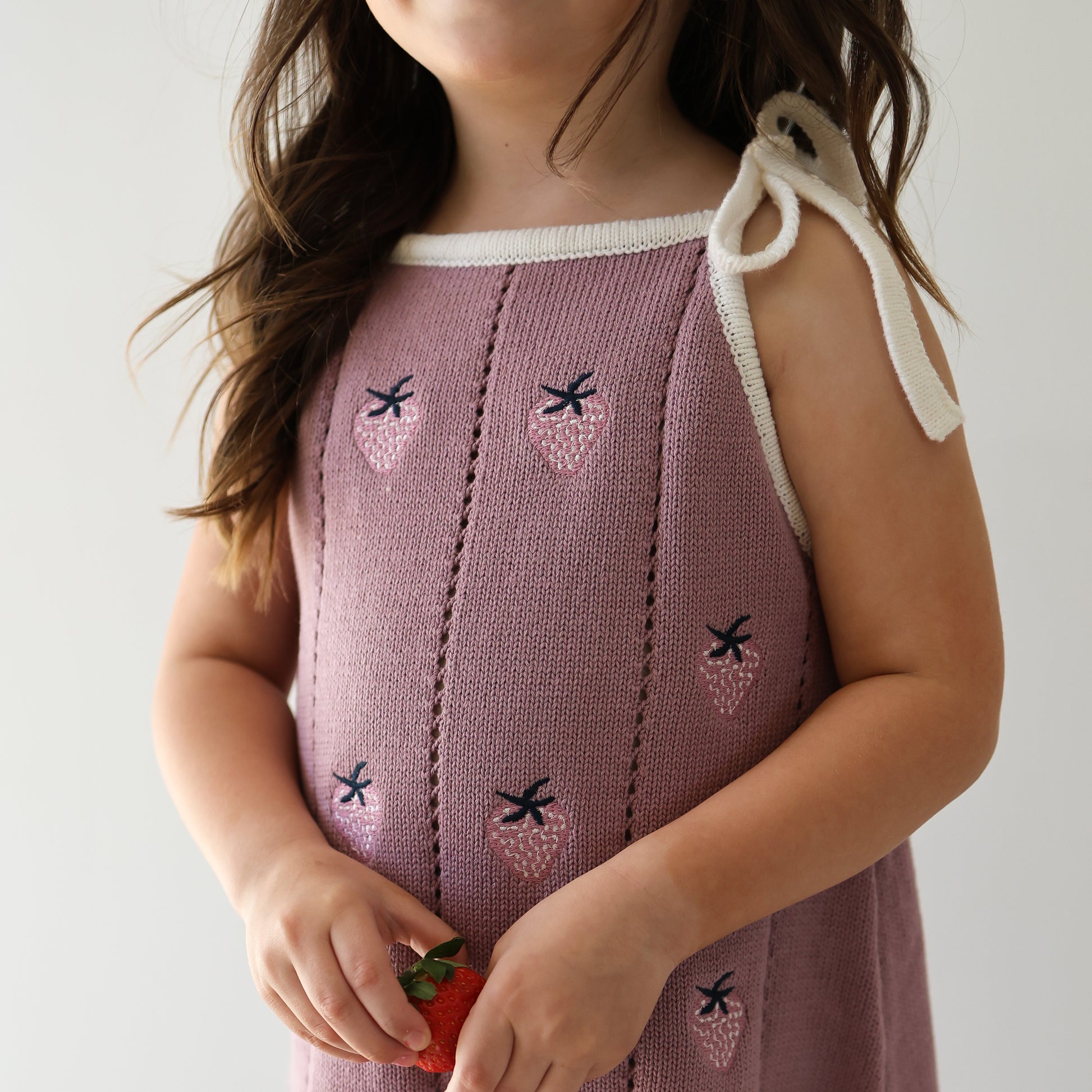 A young girl in the forever french baby Knit Dress | Matilda—a 100% cotton purple dress with white trim, strawberry prints, and adjustable tie straps—holds a fresh strawberry. Her long wavy brown hair partly covers her face.