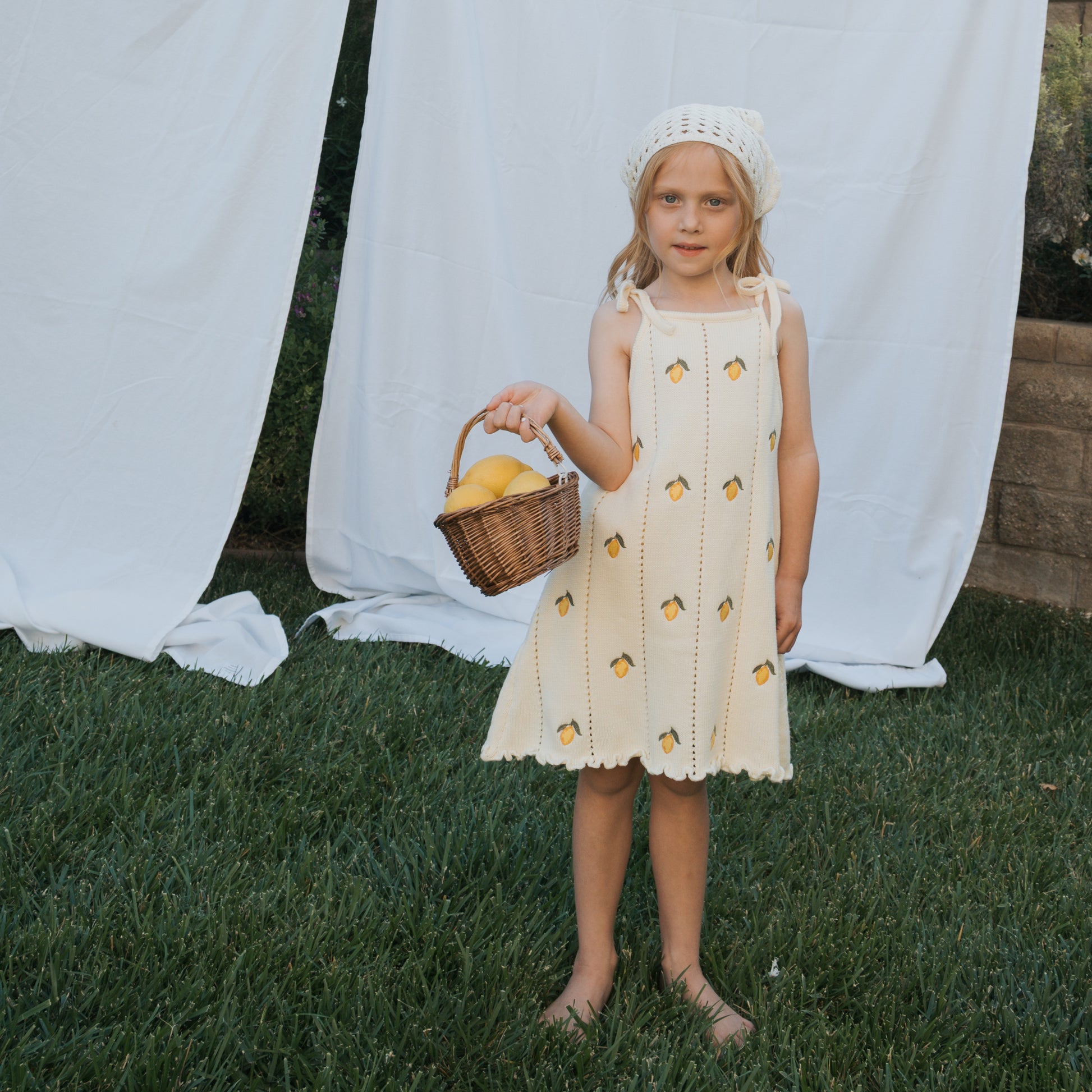 A young girl wears the forever french baby Knit Dress | Lemon and a white bonnet, standing barefoot on grass with a wicker basket of lemons. White sheets hang in the background.