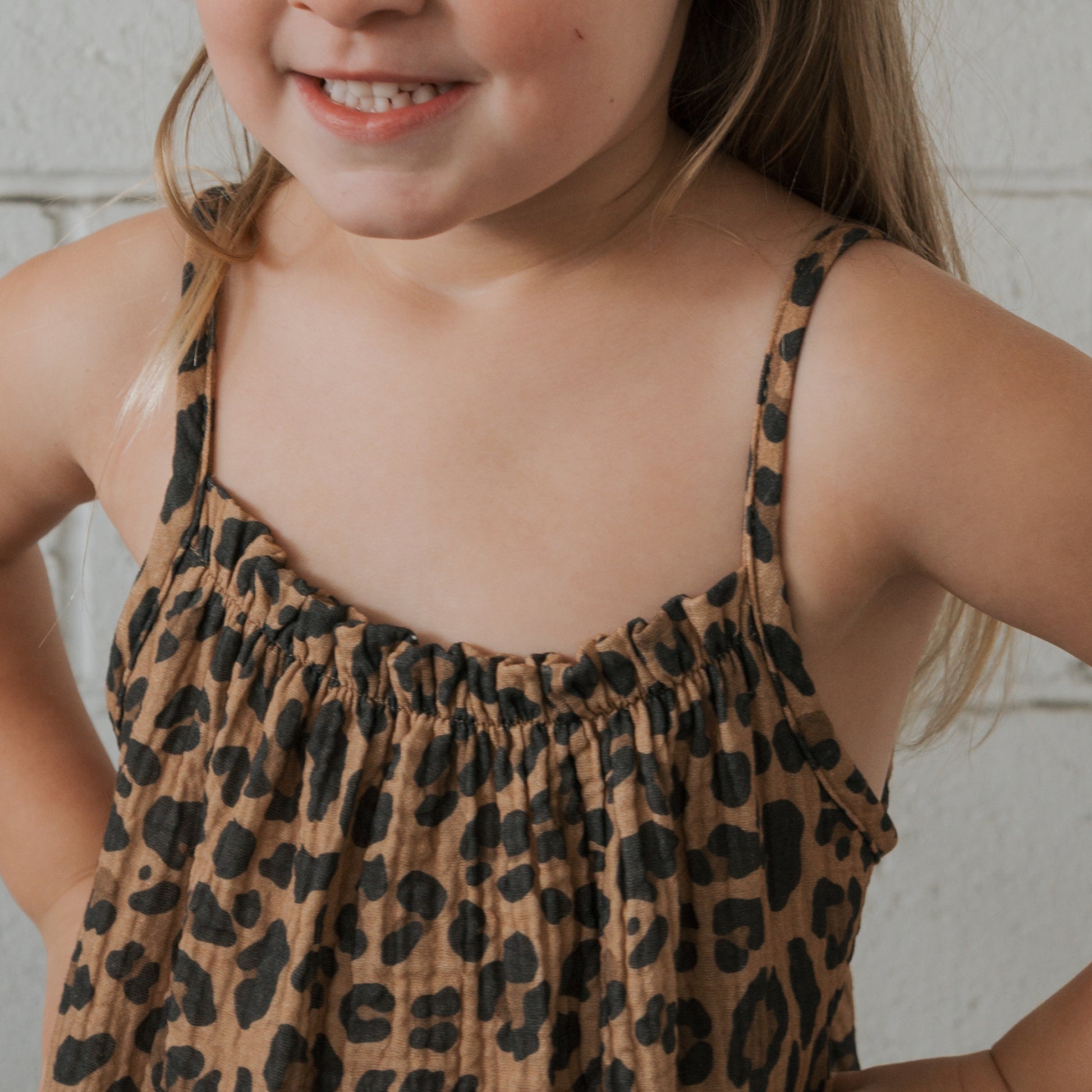 A young child smiles with hands on hips, wearing the forever french baby Child Muslin Sleeveless Dress | Leopard, featuring a black leopard print, while standing against a light-colored wall.