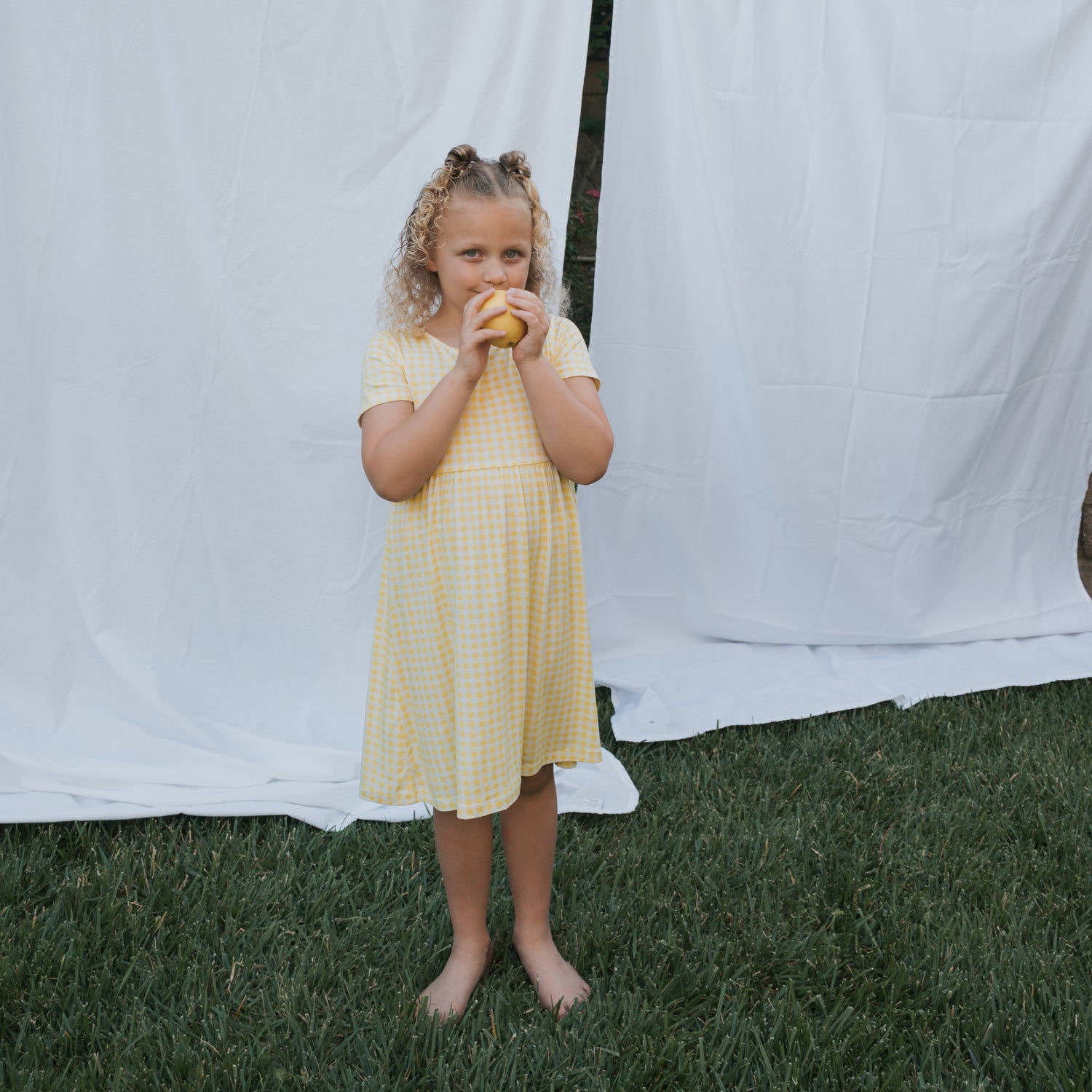 A young girl in the forever french baby Bamboo T-Shirt Dress | Citron stands barefoot on grass, holding a yellow fruit. White fabric behind her creates a soft, hypo-allergenic backdrop.