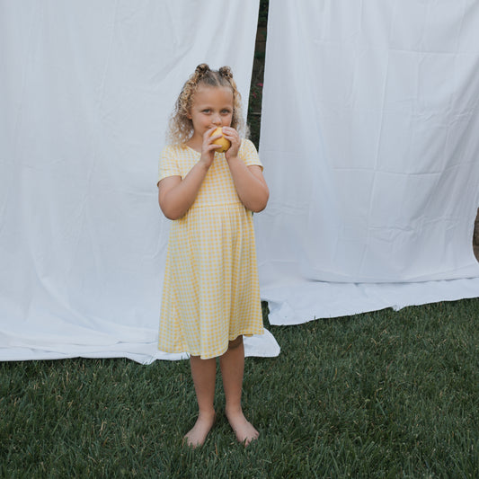 A young girl in the forever french baby Bamboo T-Shirt Dress | Citron stands barefoot on grass, holding a yellow fruit. White fabric behind her creates a soft, hypo-allergenic backdrop.