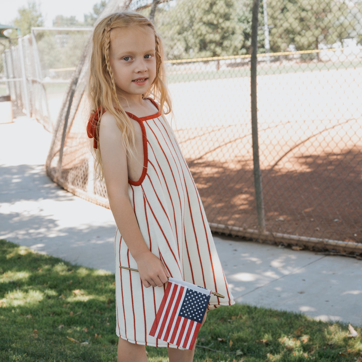 A young girl wears the forever french baby Knit Dress | Picnic Pinstripe, featuring red stripes and adjustable tie straps, as she stands on grass by a baseball field holding an American flag. Trees and a chain-link fence are behind her.