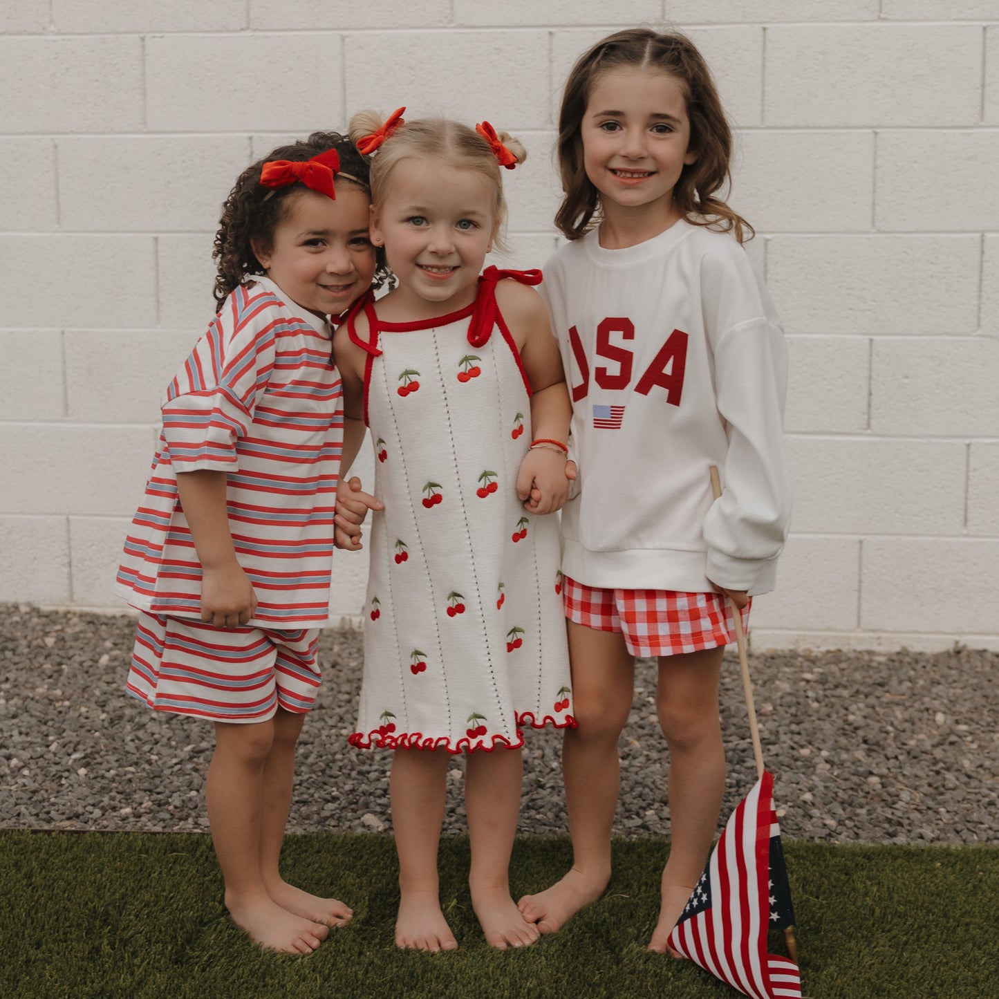 Three young girls smile outdoors. One wears the forever french baby Knit Dress | Cherry with adjustable tie straps, while another holds a small American flag. Their red, white, and blue outfits stand out against the white brick wall behind them.