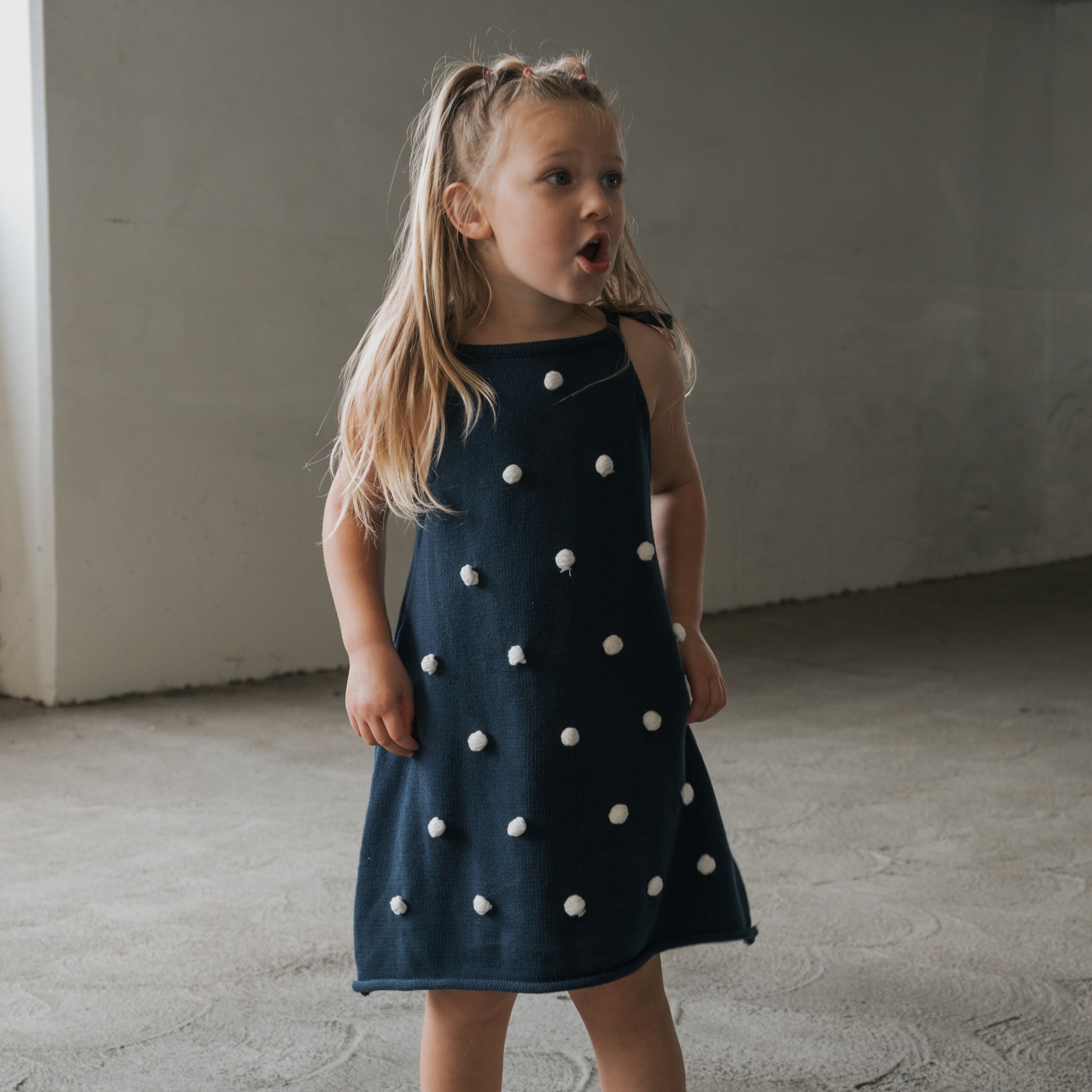A young girl with long blonde hair wears the forever french baby Knit Dress | Emery, a sleeveless navy blue 100% cotton dress with white pom-pom trim. She stands indoors on a textured concrete floor, looking to the side with her mouth open.