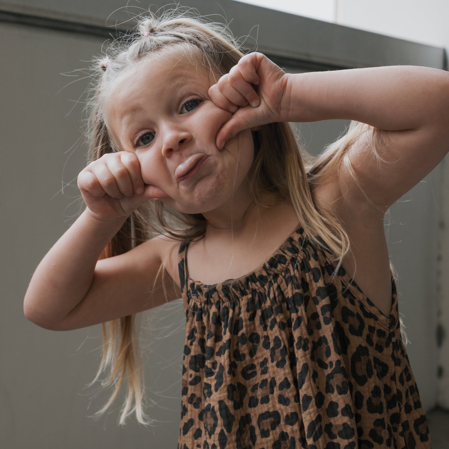 A young girl in the forever french baby Child Muslin Sleeveless Dress | Leopard makes a funny face by pulling down her mouth, standing against a plain, light background.