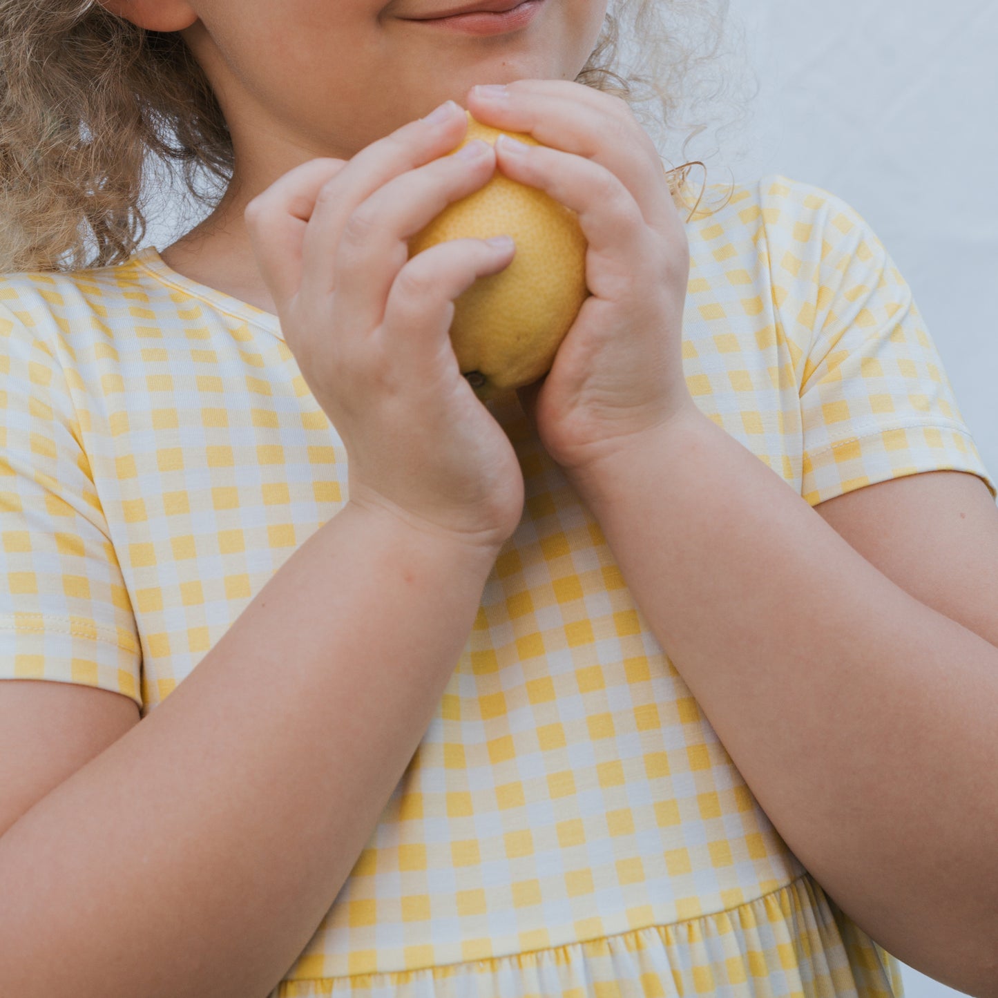 A young child wears the forever french baby Bamboo T-Shirt Dress | Citron, holding a lemon near their smile. The light background highlights the soft, hypo-allergenic fabric, with only the child's lower face and upper torso visible.