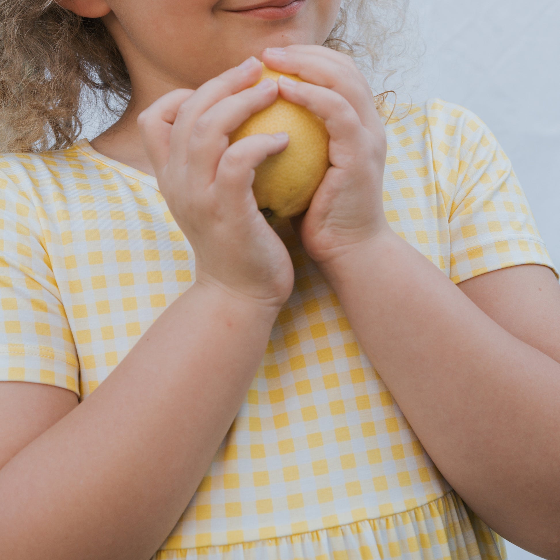 A young child wears the forever french baby Bamboo T-Shirt Dress | Citron, holding a lemon near their smile. The light background highlights the soft, hypo-allergenic fabric, with only the child's lower face and upper torso visible.