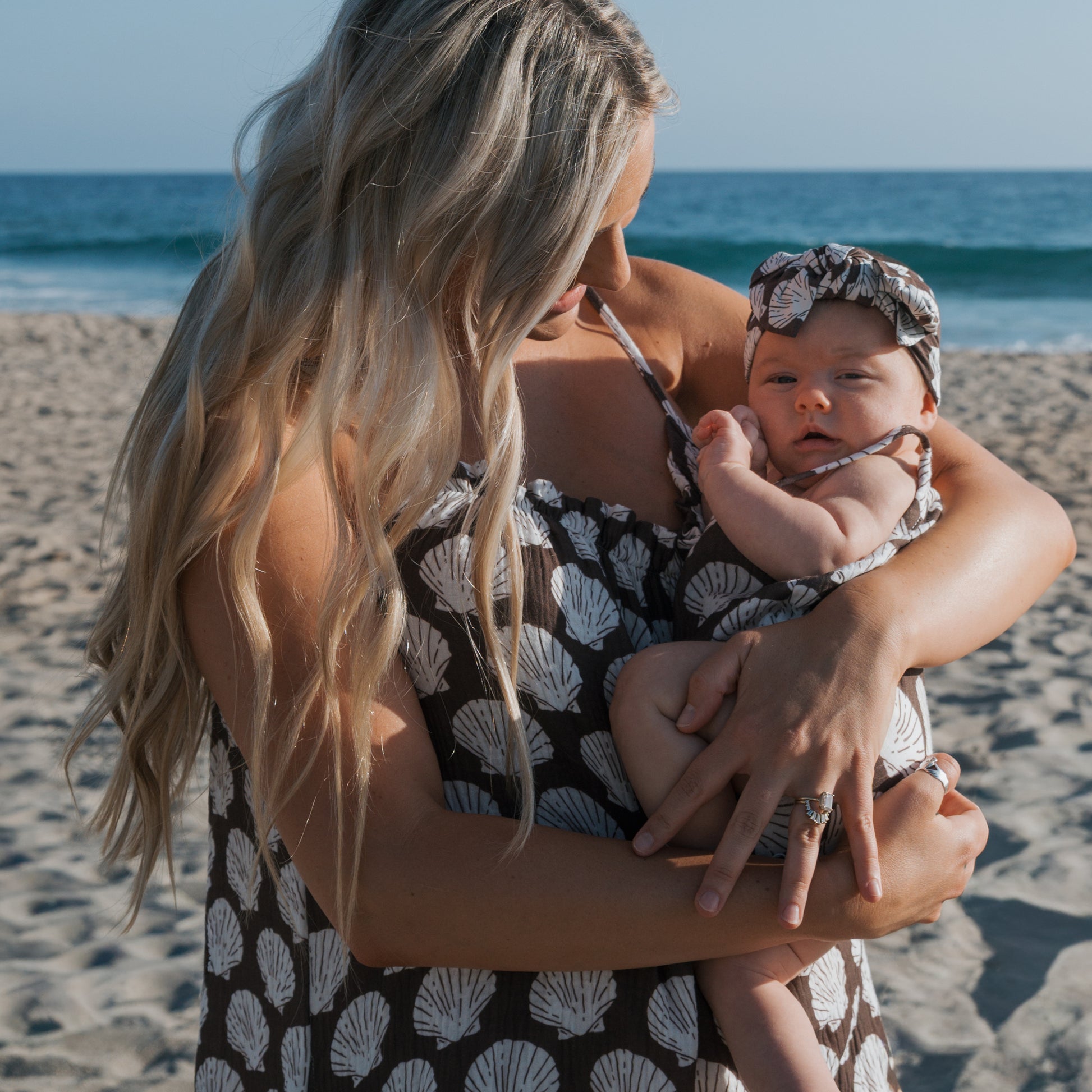 A woman with long blonde hair holds a baby at the beach in matching outfits. The baby wears the forever french baby Muslin Sleeveless Tank Romper | Seashells, made of 100% cotton with white shell patterns. Ocean and sand appear under a clear sky.
