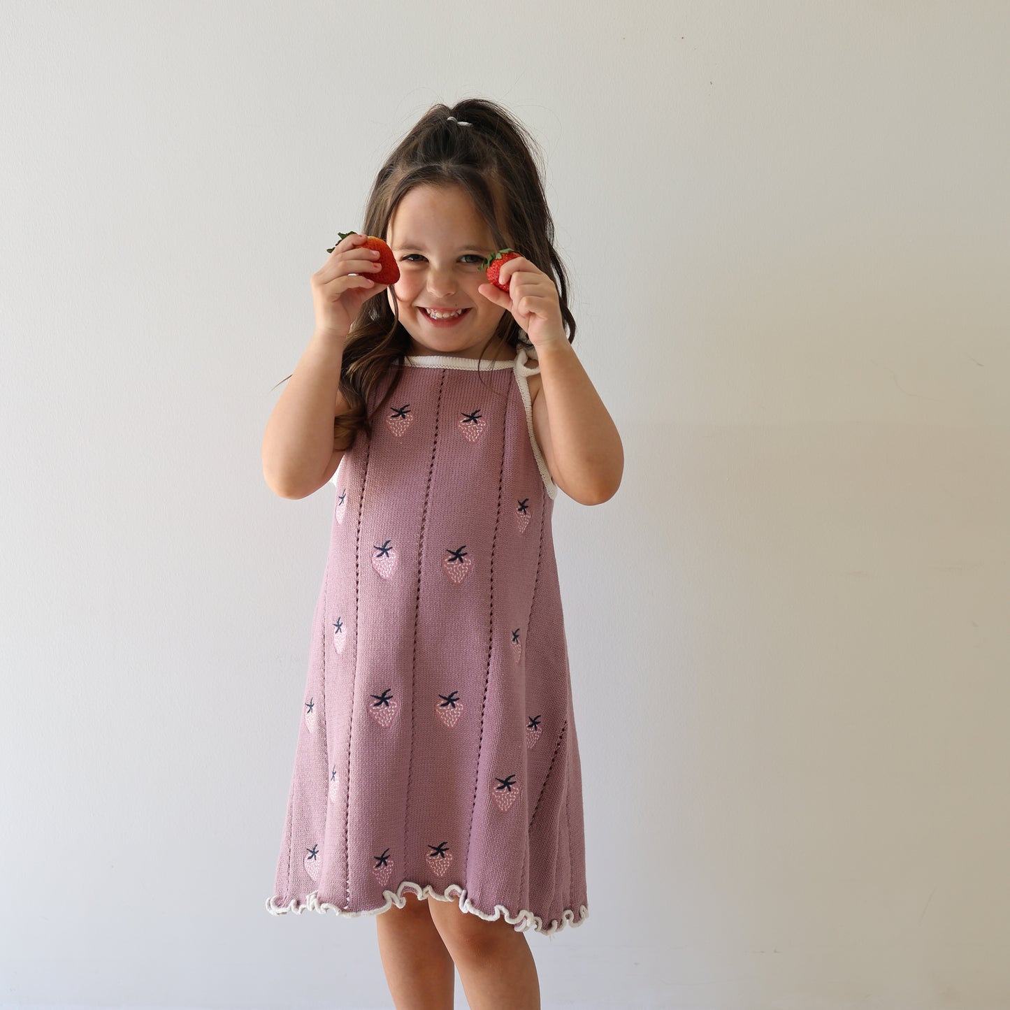 A young girl smiles while holding two strawberries to her eyes, wearing the forever french baby Knit Dress | Matilda—a pink 100% cotton knit dress with a scalloped hem and adjustable tie straps—against a plain white background.