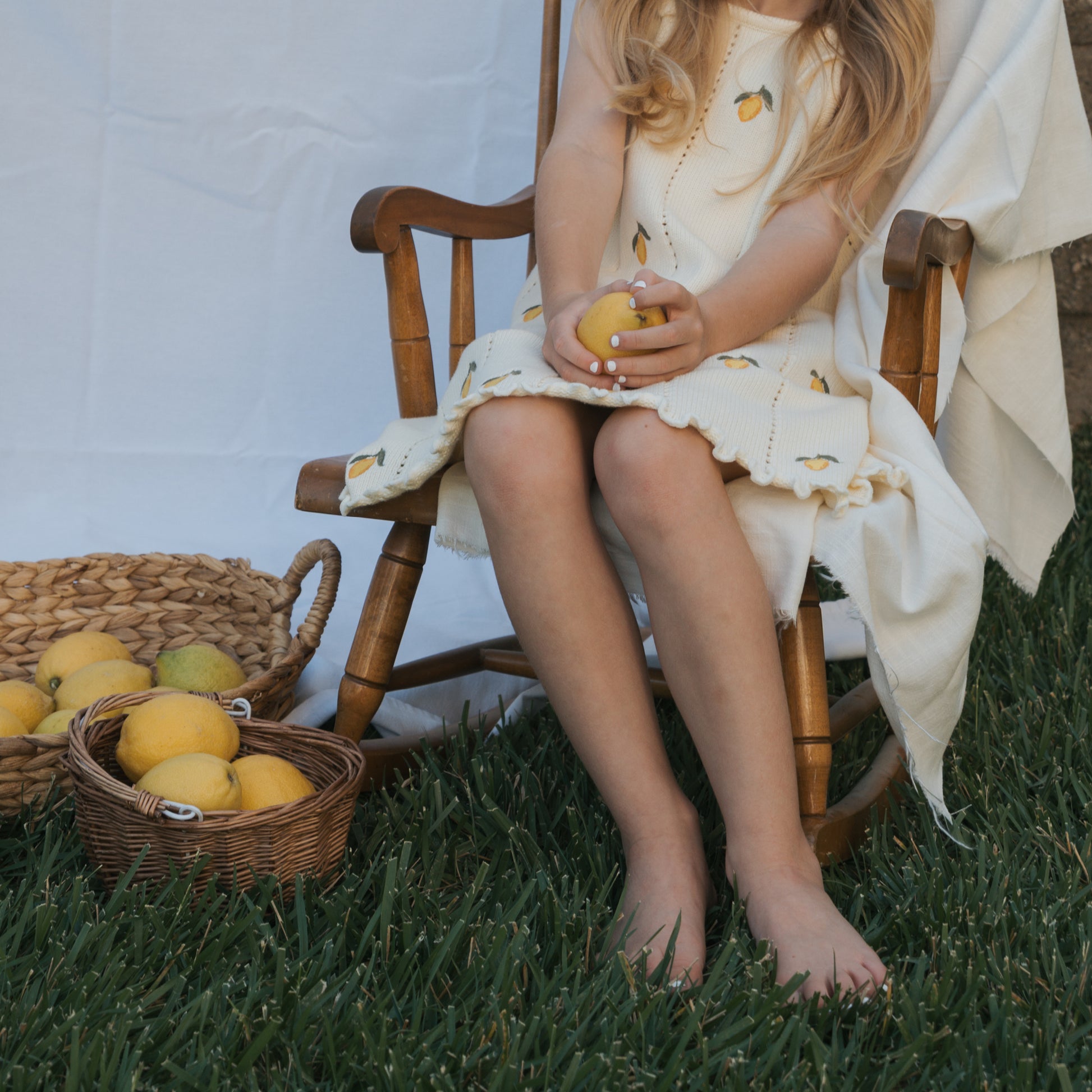 A young girl in the forever french baby Knit Dress | Lemon, made from 100% cotton with adjustable tie straps, sits barefoot on a wooden chair outdoors holding a lemon. Two wicker baskets of lemons rest on the grass nearby.