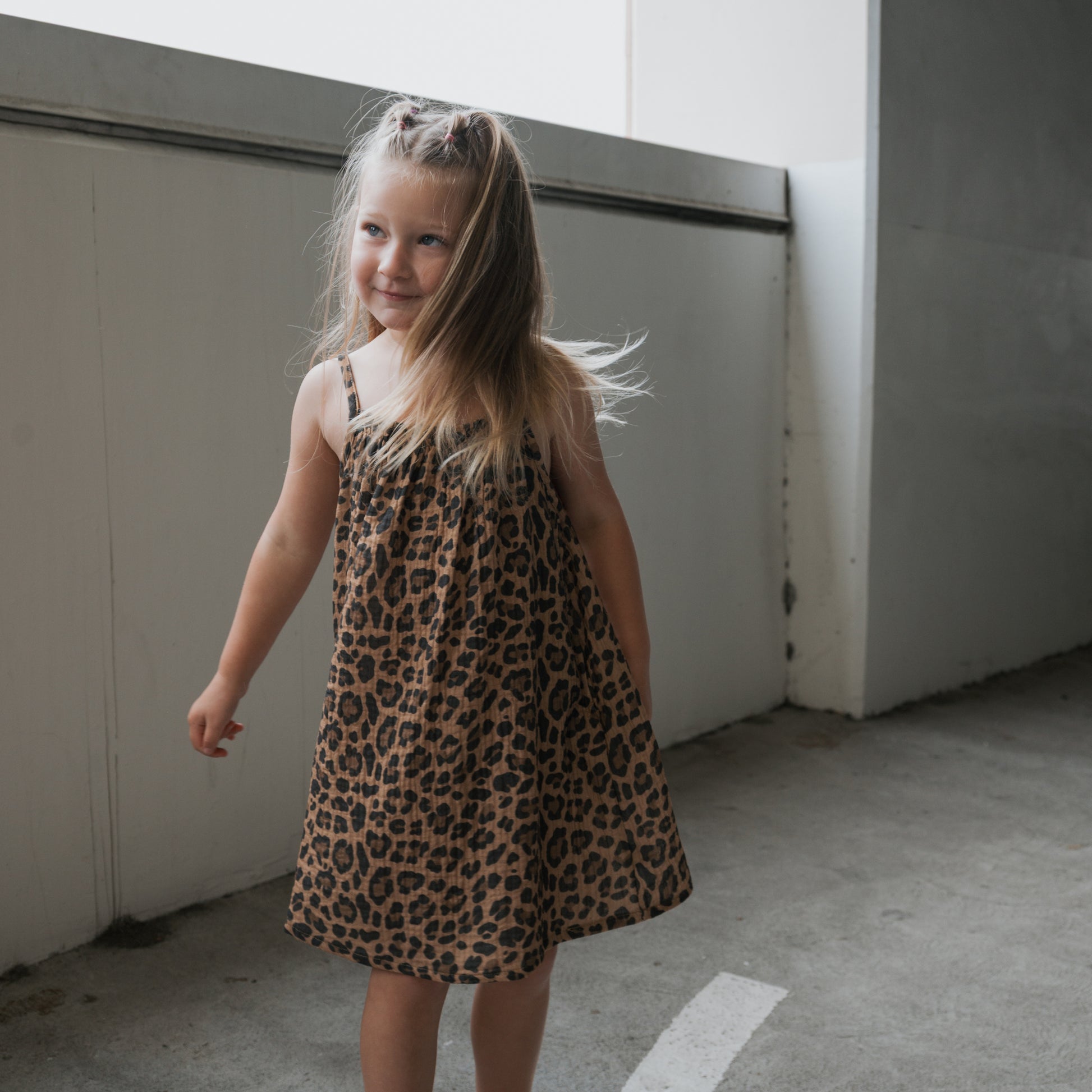 A young girl with long blonde hair smiles while wearing the forever french baby Child Muslin Sleeveless Dress in Leopard print, standing on a concrete floor in a bright, modern indoor setting.