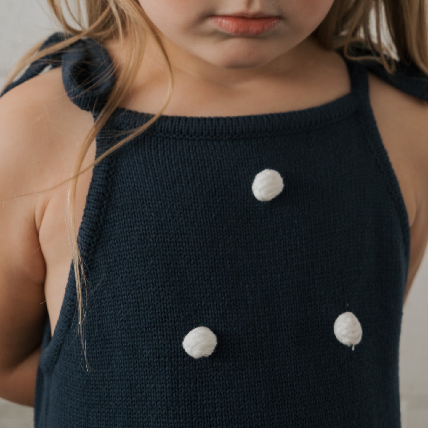 A young child wears the forever french baby Knit Dress | Emery—navy blue, 100% cotton, sleeveless with adjustable tie straps and three white pom-poms—standing against a neutral background.