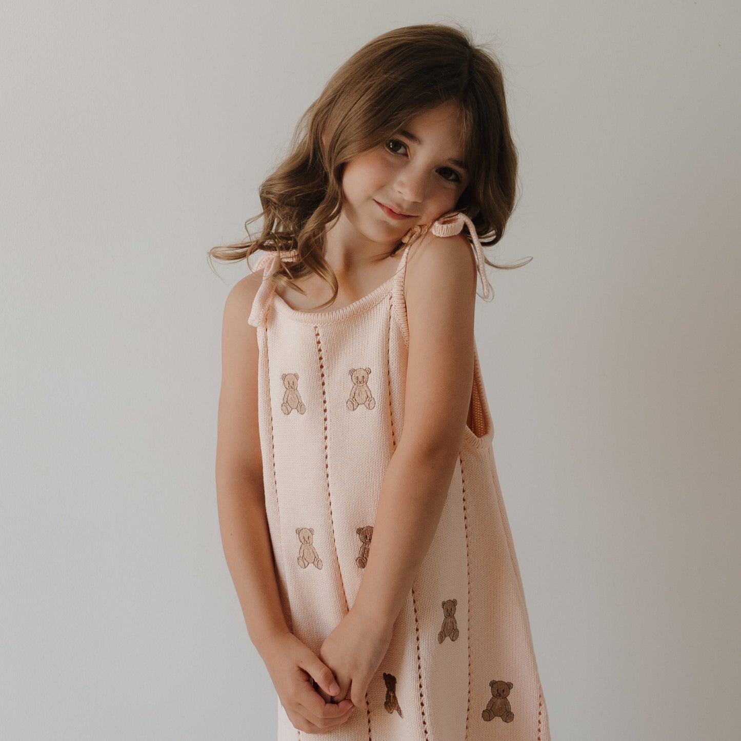 A young girl with wavy brown hair smiles softly, tilting her head as she stands against a plain light wall, wearing the forever french baby Knit Dress | Lottie—crafted from 100% cotton knit in light pink with an embroidered teddy bear pattern.