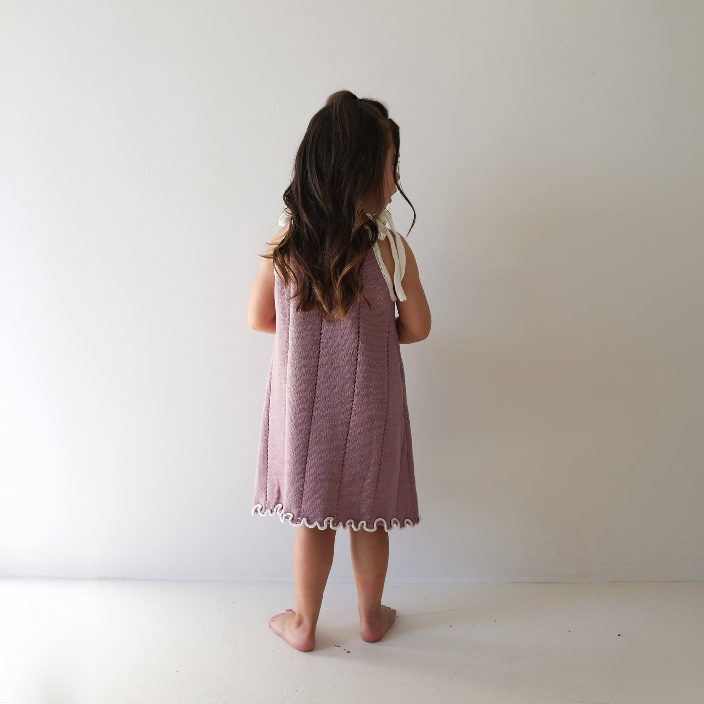A young child with long brown hair stands barefoot, facing away from the camera, wearing the forever french baby Knit Dress | Matilda— a sleeveless mauve 100% cotton knit dress with white trim and adjustable tie straps.