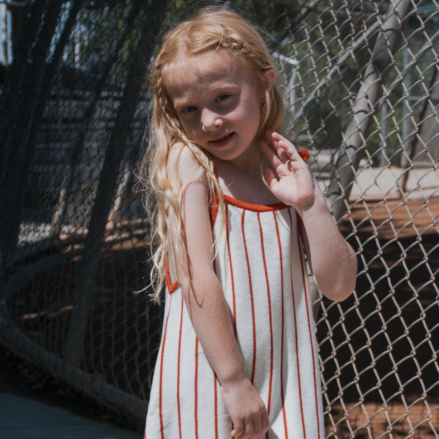 A young girl with long blonde hair in braids wears the forever french baby Knit Dress | Picnic Pinstripe, a sleeveless, 100% cotton white dress with red stripes. She smiles and raises her hand by a chain-link fence on a sunny day.