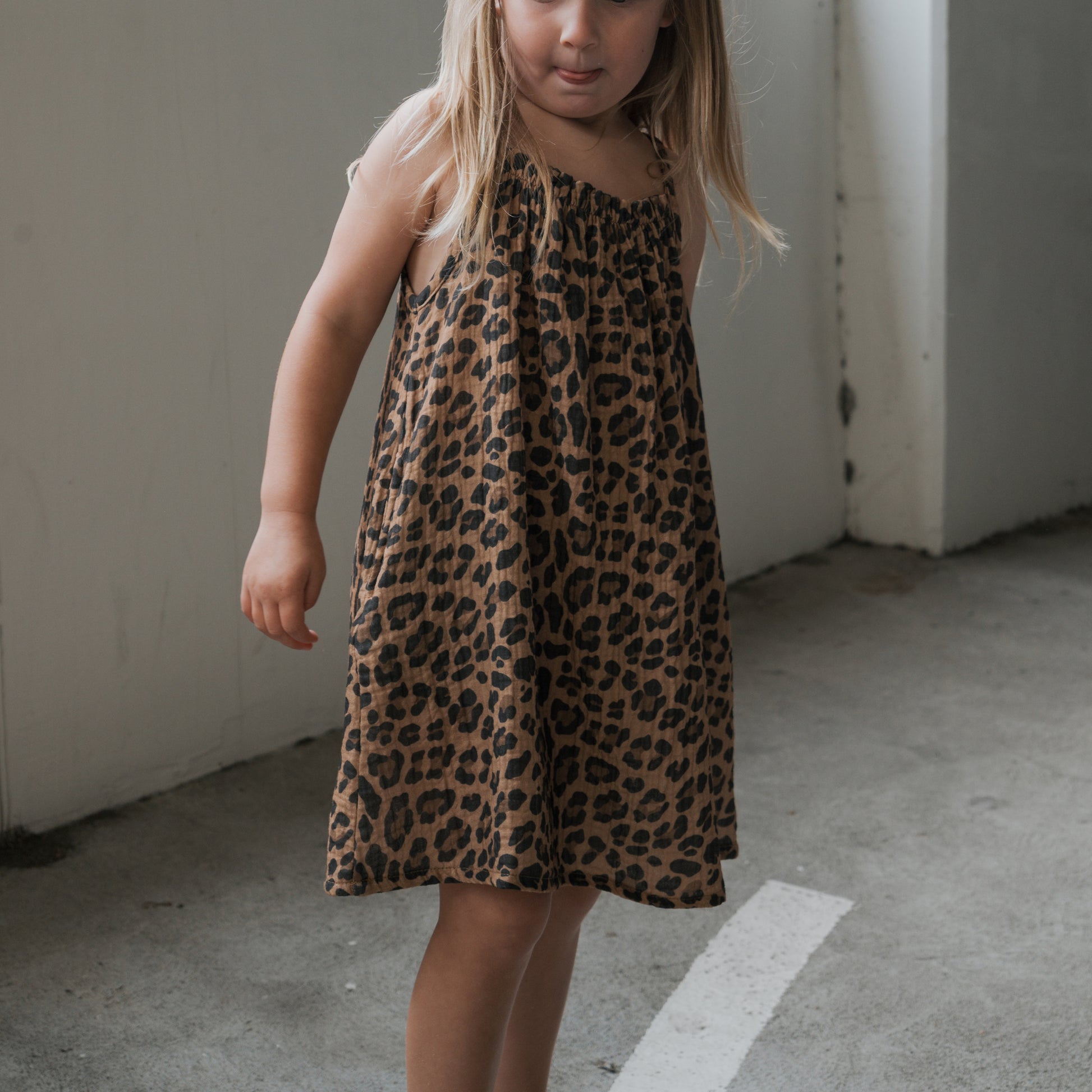 A young girl with long blonde hair wears the forever french baby Child Muslin Sleeveless Dress in Leopard, standing indoors on a concrete floor and gazing slightly to the side.