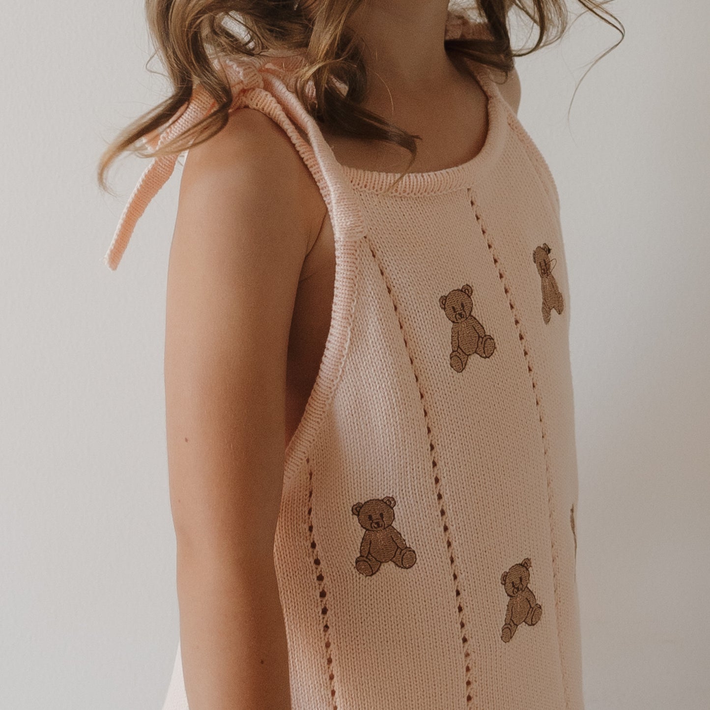 A child wears the forever french baby Knit Dress | Lottie, a light pink 100% cotton dress with tied shoulder straps and small embroidered teddy bears, standing against a plain white background.