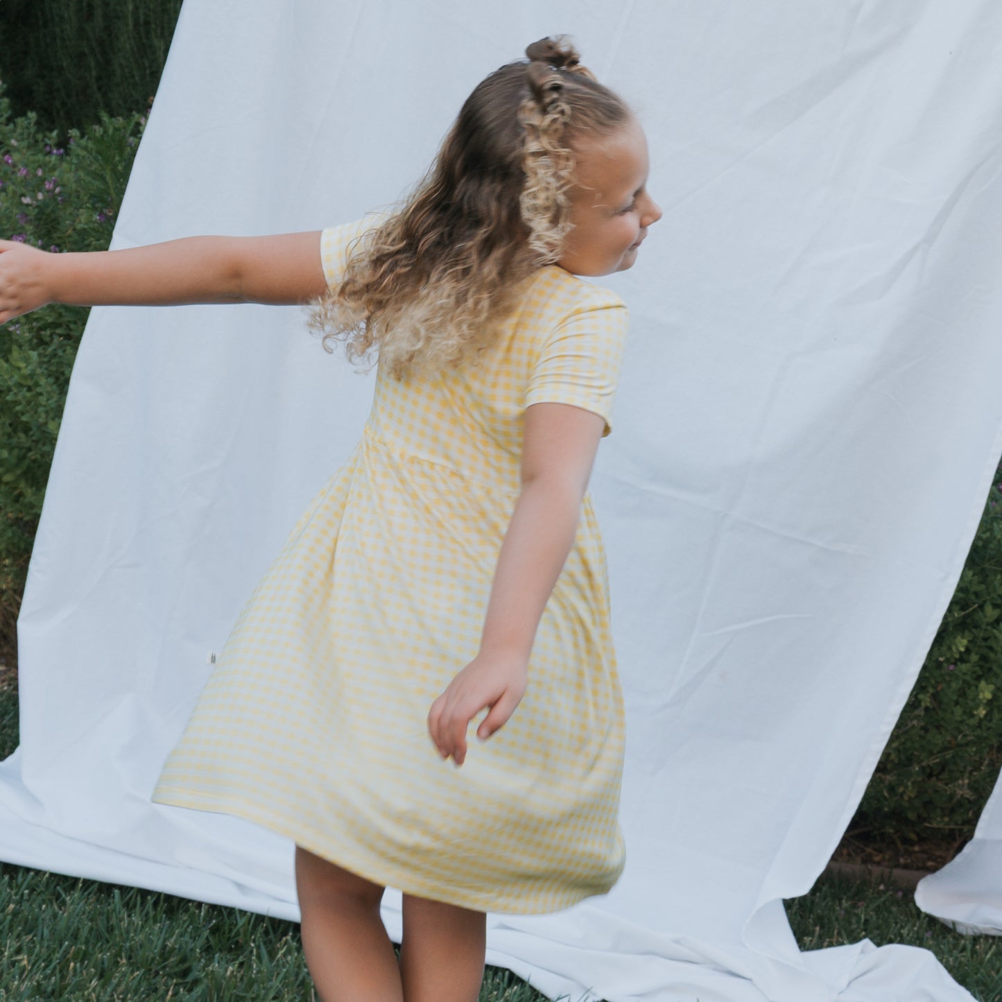 A young girl with curly hair twirls on the grass wearing the Bamboo T-Shirt Dress | Citron by forever french baby, with a white sheet hanging in the background.