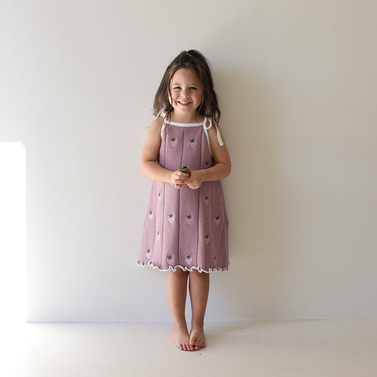 A young girl with long brown hair stands barefoot against a white wall, smiling. She wears the Knit Dress | Matilda by forever french baby—a pink 100% cotton dress with white trim, flower patterns, and adjustable tie straps—holding a small object.