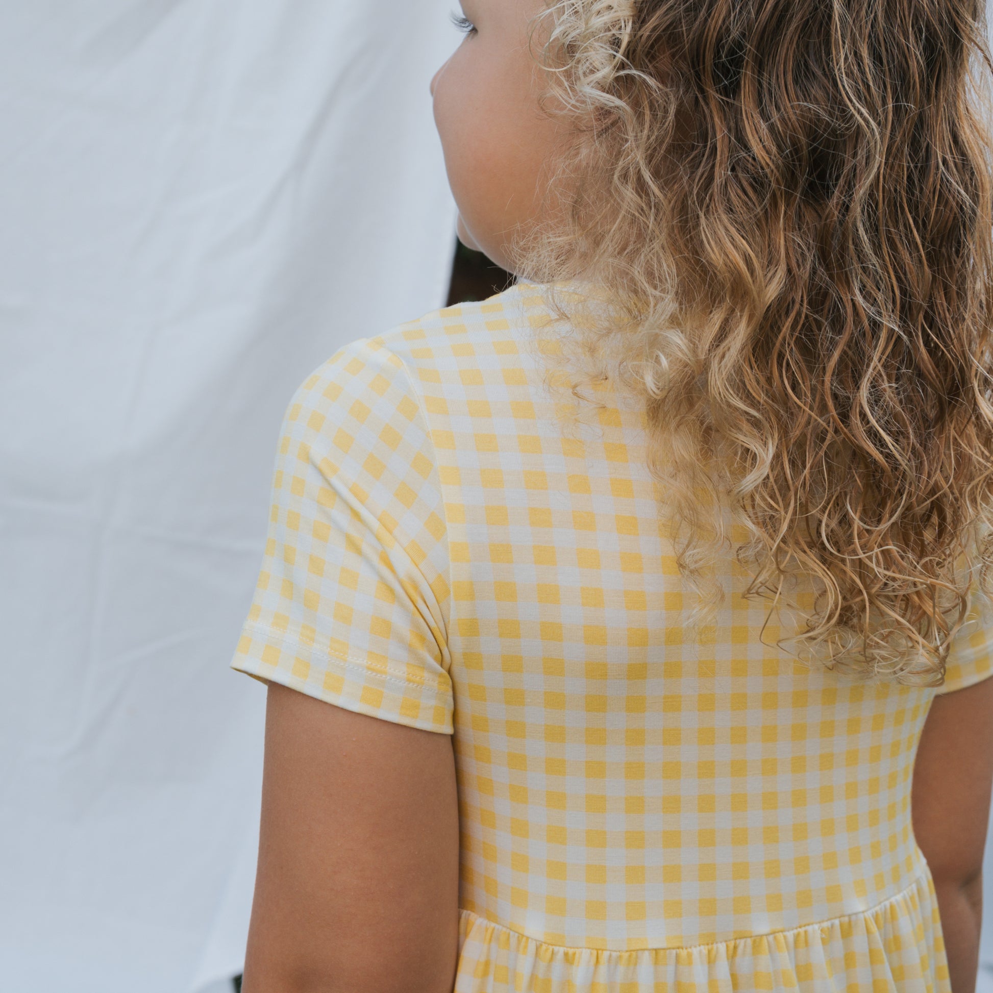 A young child with curly hair stands facing away from the camera in a forever french baby Bamboo T-Shirt Dress | Citron, featuring yellow and white checks—blending style with hypo-allergenic comfort against a white background.