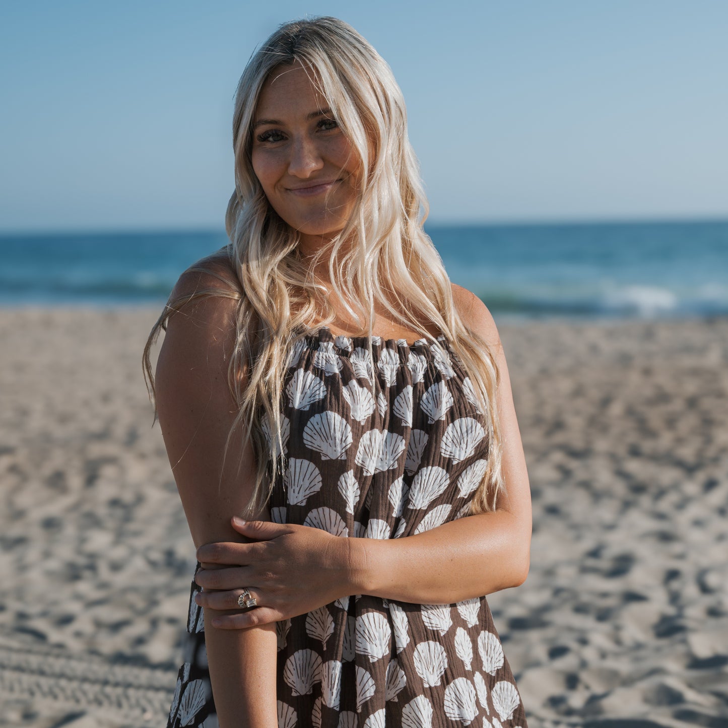 A woman with long blonde hair wears the forever french baby Women's Muslin Sleeveless Dress | Seashells, smiling gently as she stands on a sandy beach with the ocean and blue sky in the background.