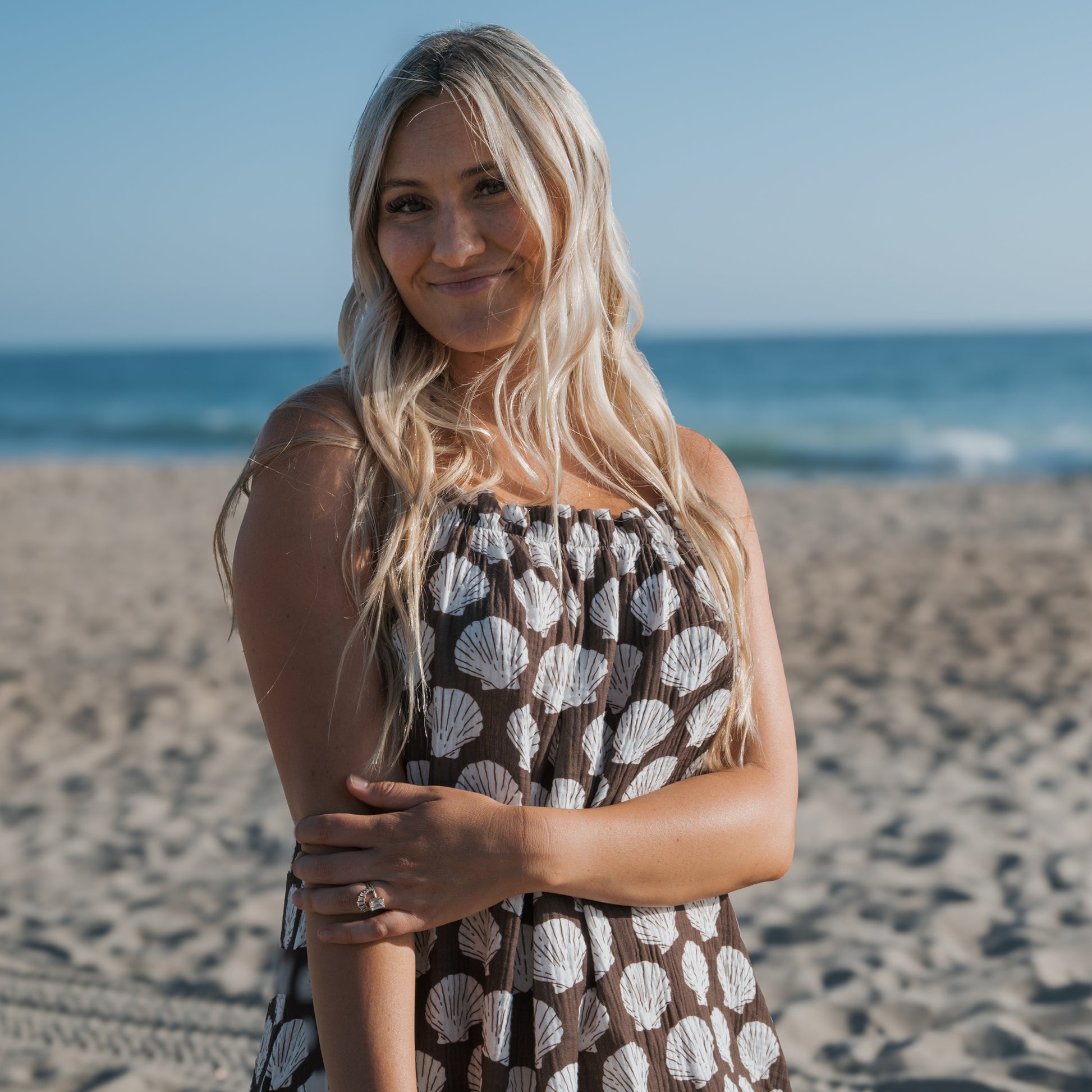 A woman with long blonde hair wears the forever french baby Women's Muslin Sleeveless Dress | Seashells, smiling gently as she stands on a sandy beach with the ocean and blue sky in the background.