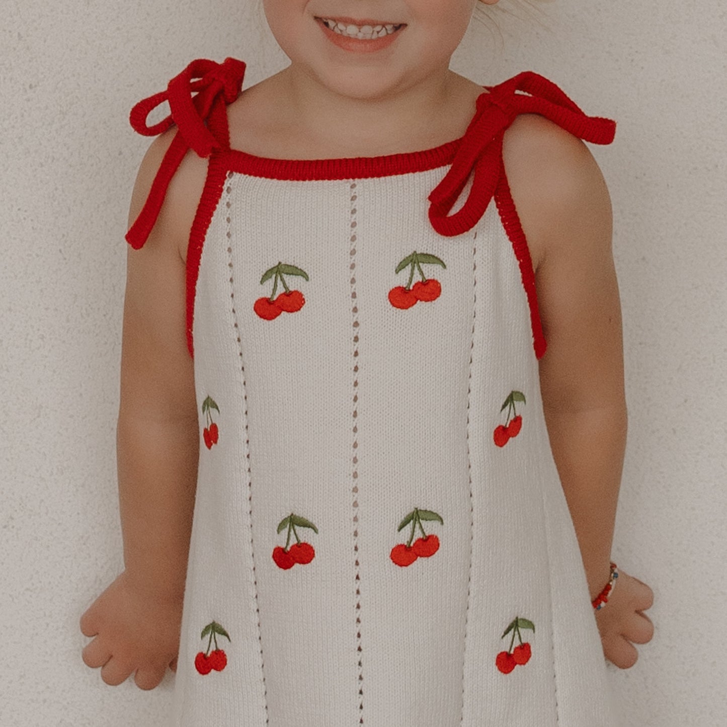 A young child smiles in the forever french baby Knit Dress | Cherry, a sleeveless white knit dress with red cherries, red trim, and adjustable tie shoulder straps, set against a plain light background.