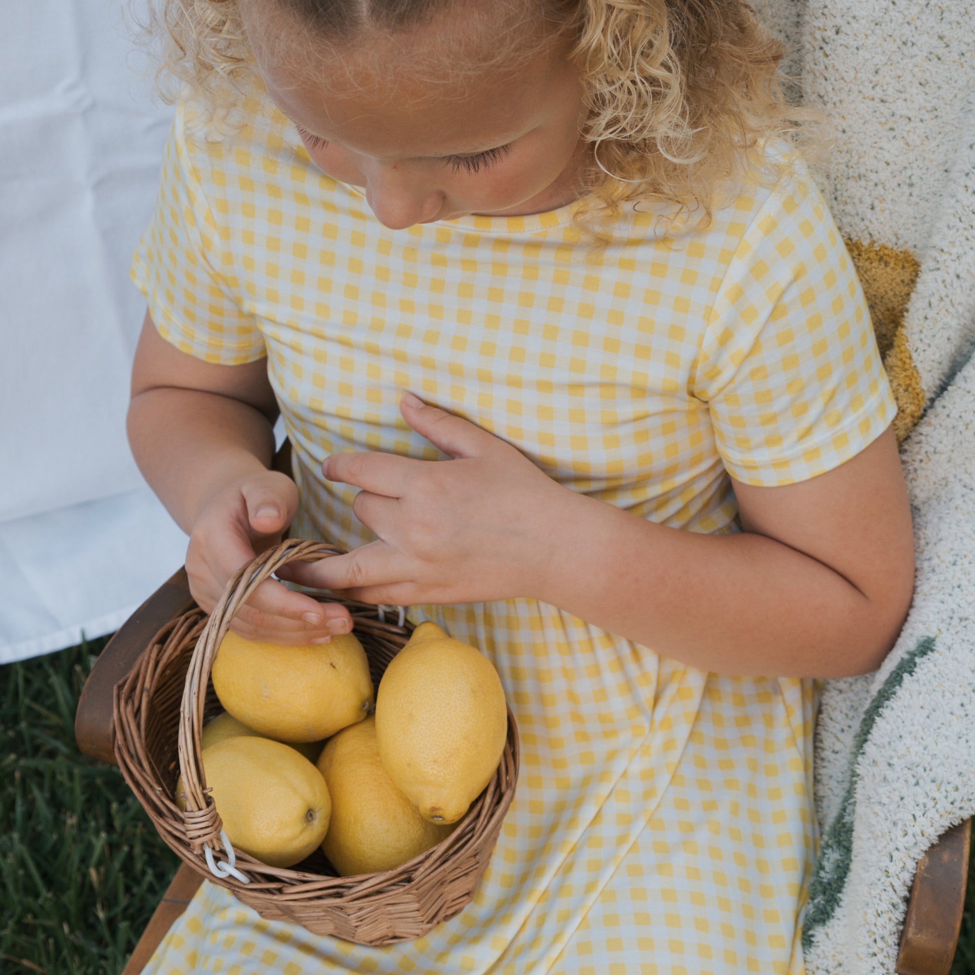 A young child with curly blonde hair sits outdoors, viewed from above, holding a wicker basket of lemons while wearing the hypo-allergenic Bamboo T-Shirt Dress | Citron from forever french baby.