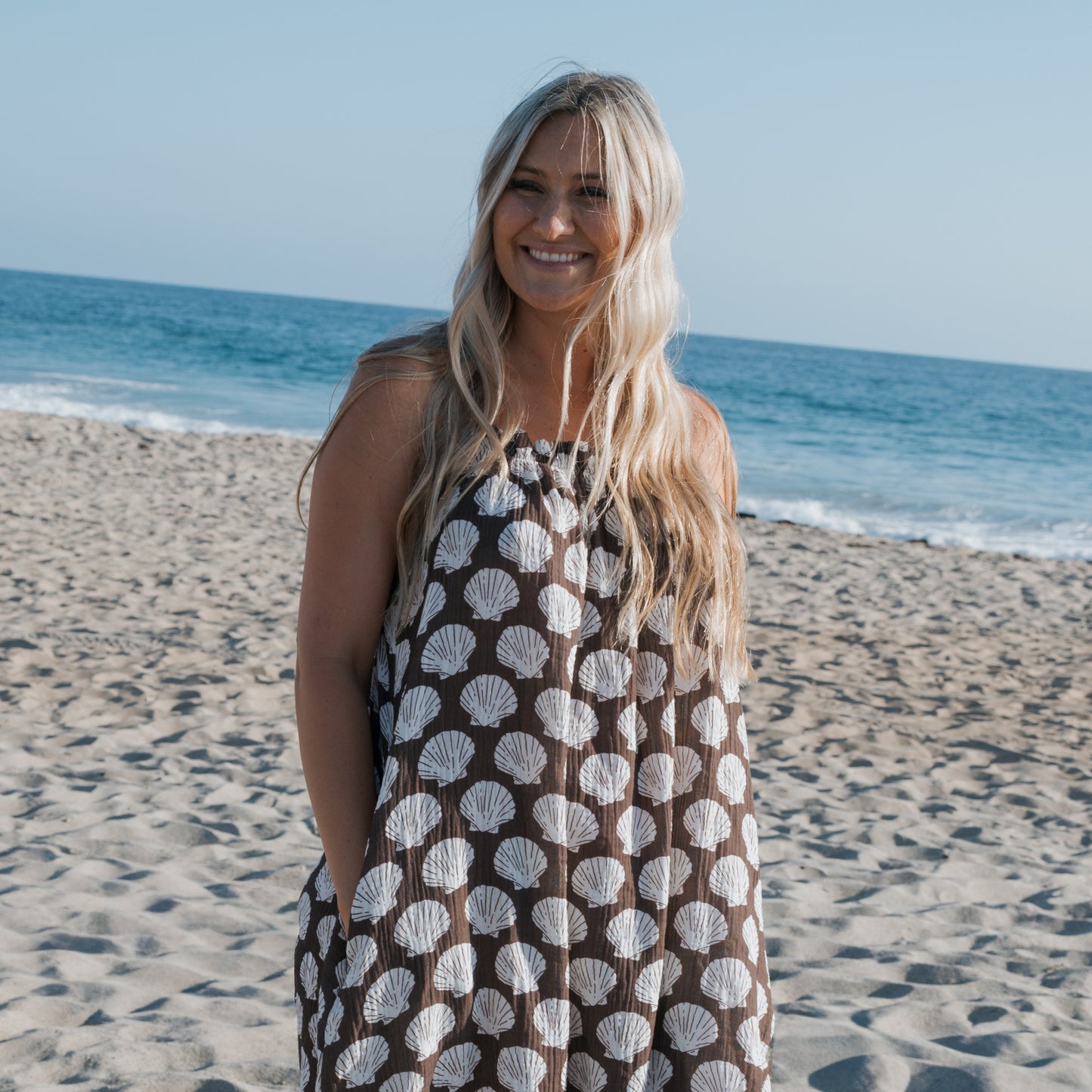 A woman with long blonde hair smiles on a sandy beach, wearing the forever french baby Women's Muslin Sleeveless Dress in Seashells, with the ocean and blue sky in the background.