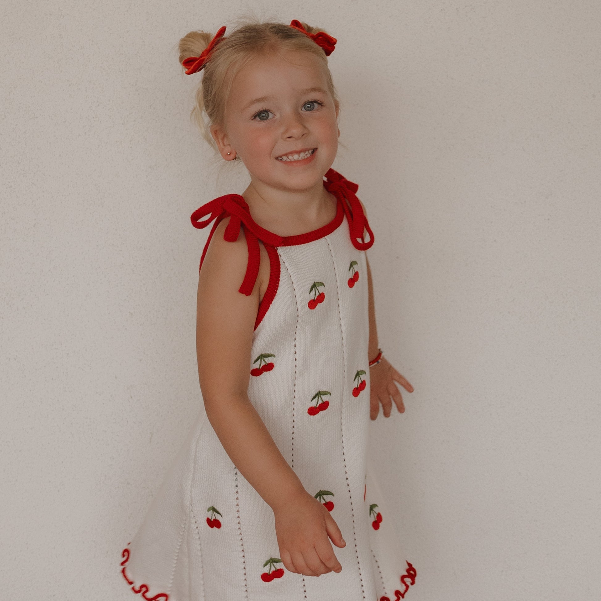 A smiling young girl with blonde pigtails tied with red bows wears the forever french baby Knit Dress | Cherry, a cute 100% cotton white dress with cherry patterns and red trim, standing before a plain light-colored wall.