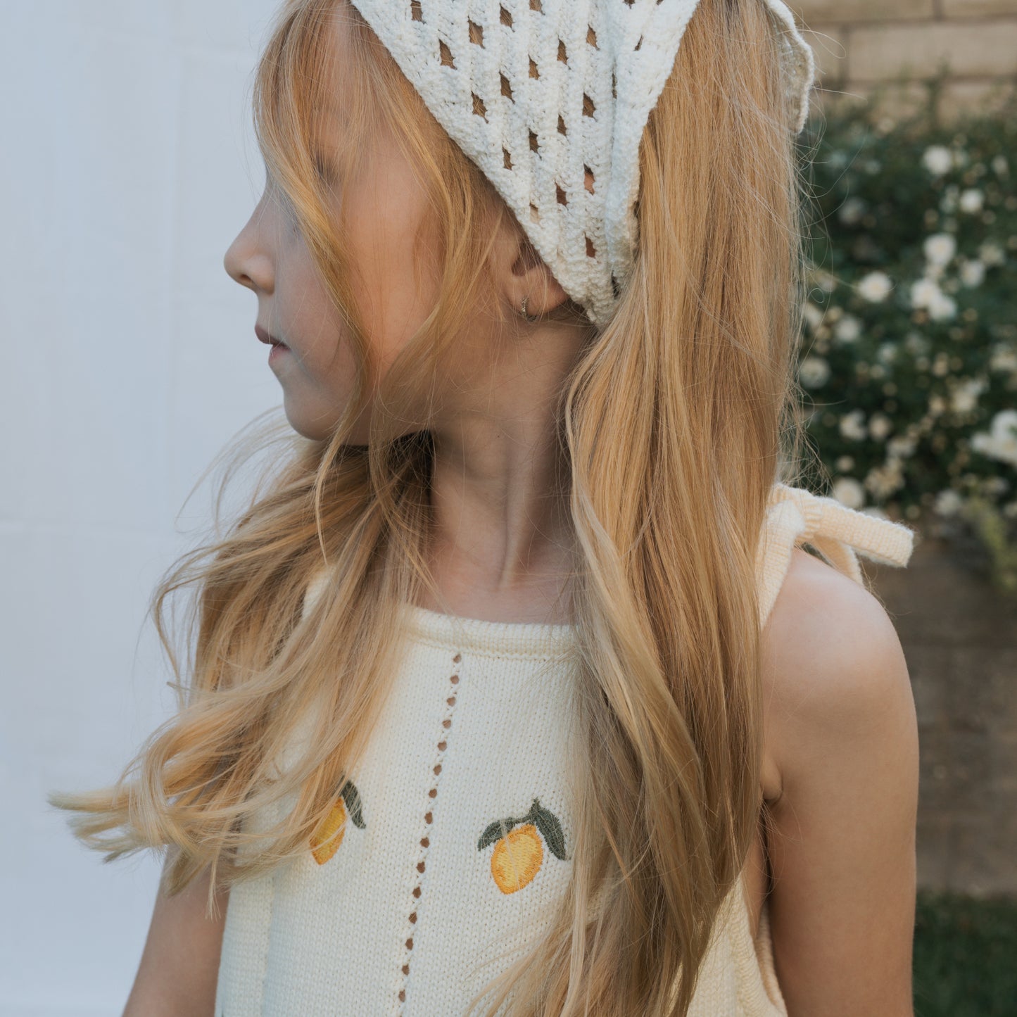 A young girl with long blonde hair wears the forever french baby Knit Dress | Lemon, a sleeveless 100% cotton dress with lemon print and adjustable tie straps, looking to the side against a blurred garden background.