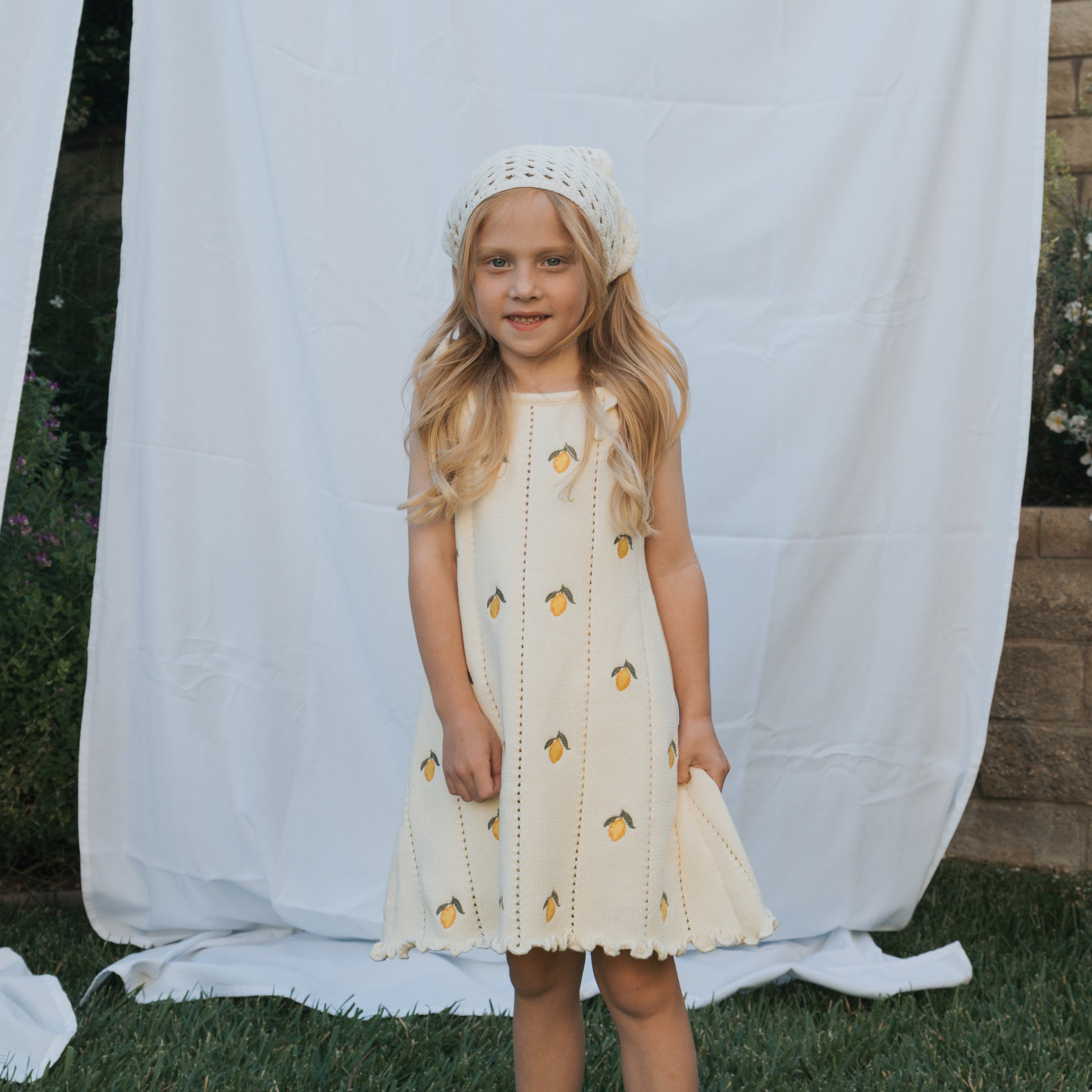 A young girl with long blonde hair wears the forever french baby Knit Dress | Lemon, featuring adjustable tie straps and yellow lemon patterns, standing on grass before a white fabric backdrop outdoors.