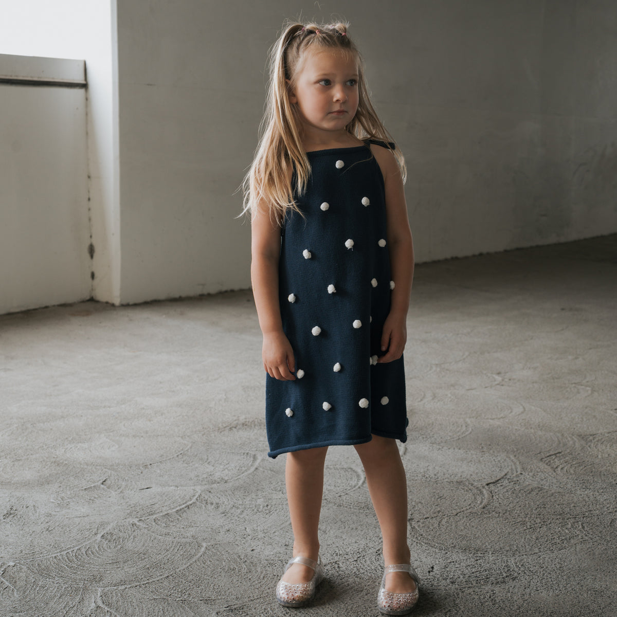 A young girl with long blonde hair stands indoors on a textured floor, wearing the forever french baby Knit Dress | Emery—navy blue 100% cotton with adjustable tie straps and white pom-poms—and sparkly shoes, looking slightly to the side.