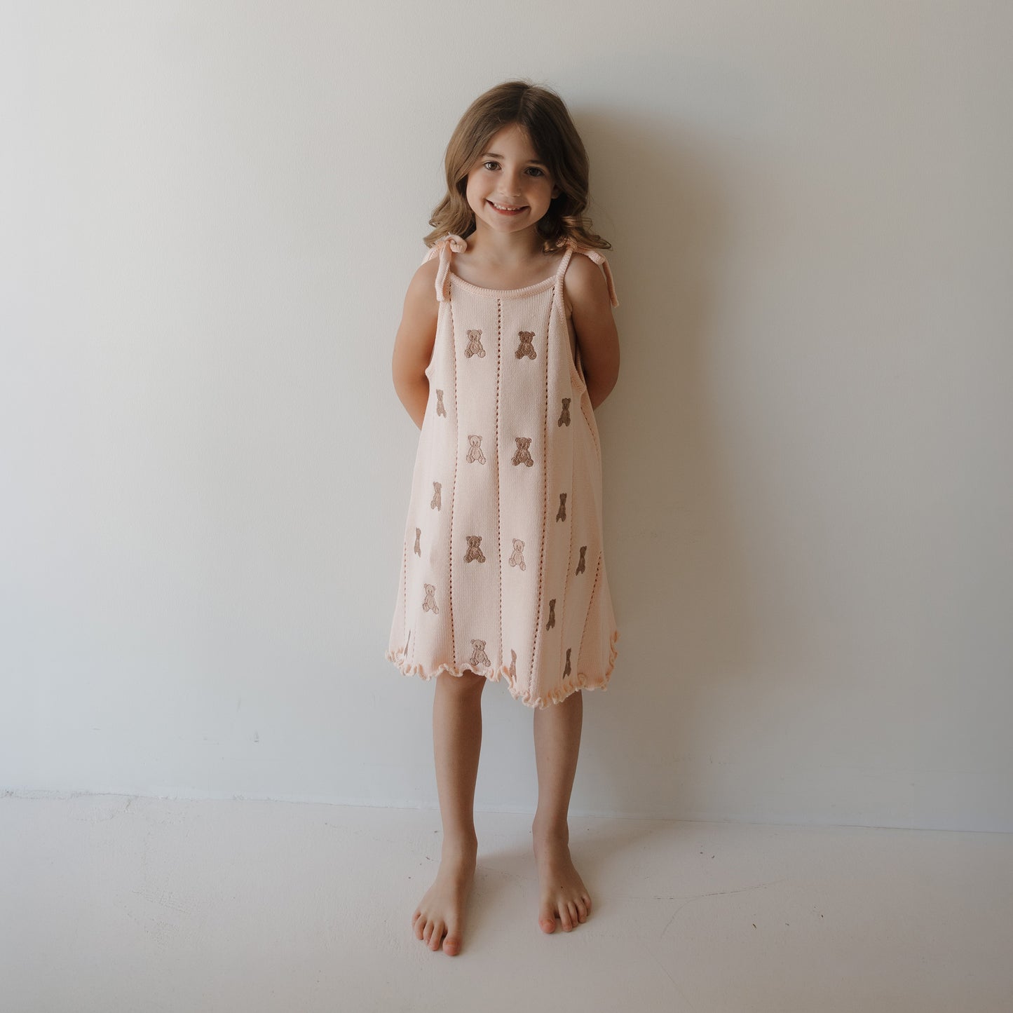 A young girl with brown hair stands barefoot against a white wall, smiling and wearing the forever french baby Knit Dress | Lottie—a light pink sleeveless 100% cotton dress featuring an embroidered teddy bear pattern.