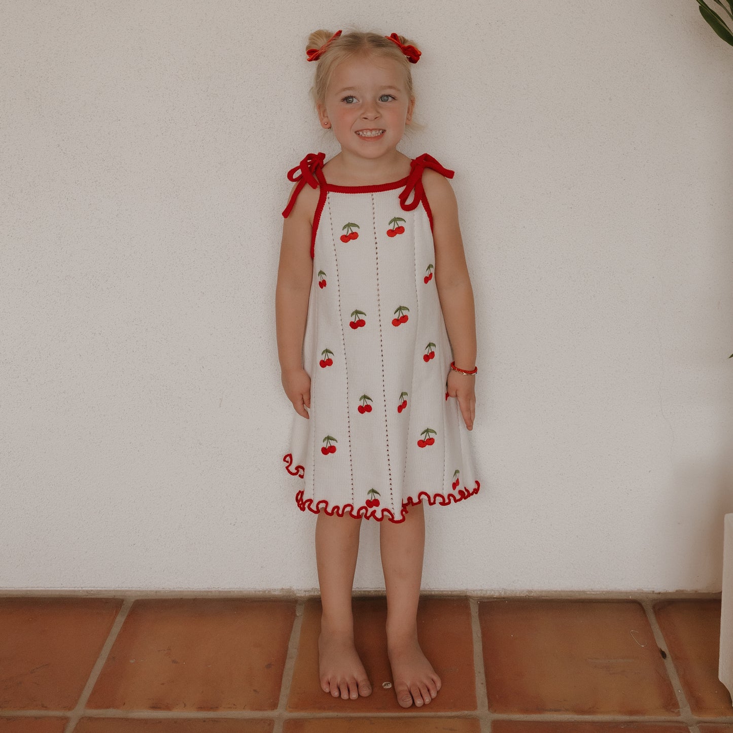 A young girl with blonde pigtails and red bows stands barefoot on terracotta tiles, smiling against a white wall. She wears the forever french baby Knit Dress | Cherry, a white 100% cotton dress with red trim and cherry prints.
