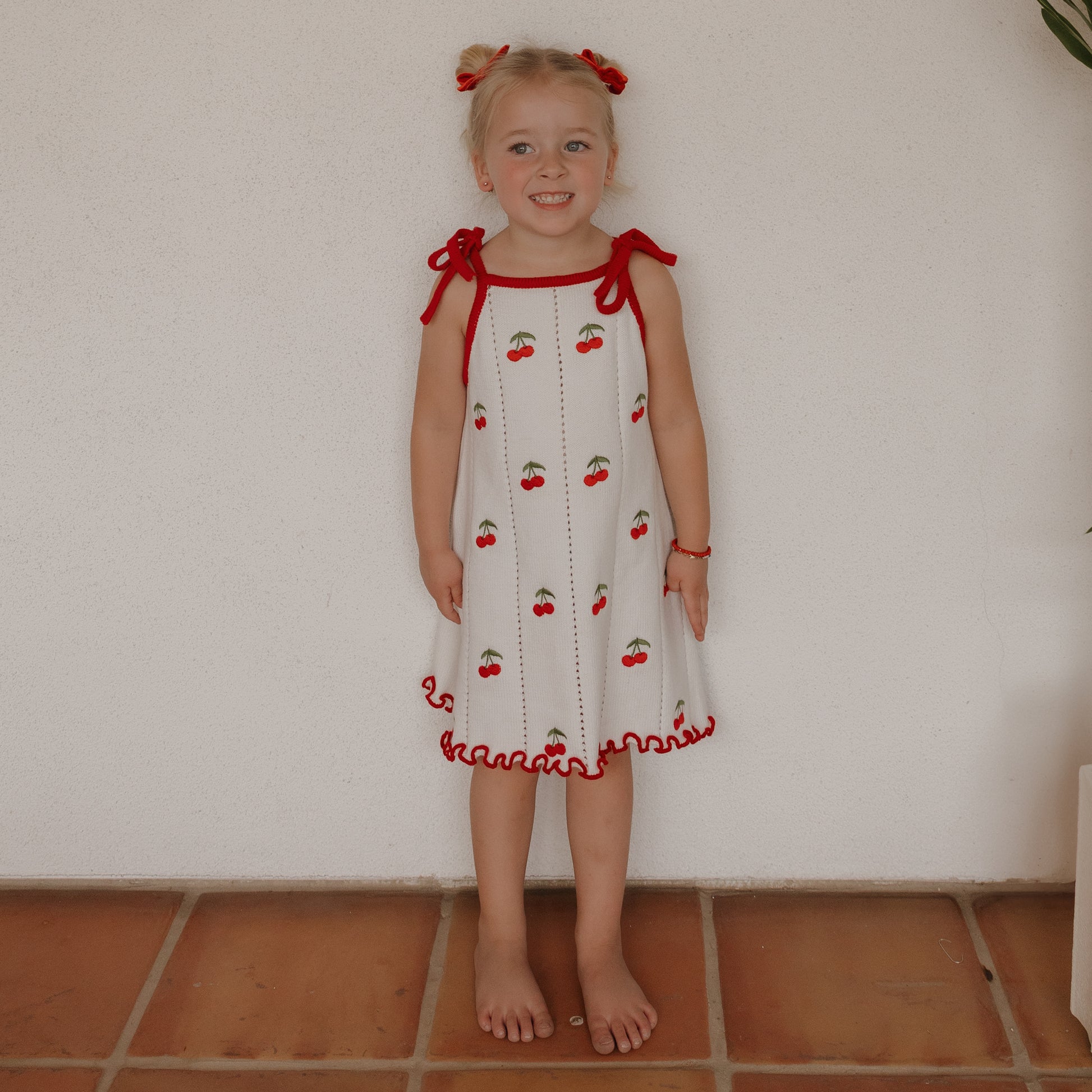 A young girl with blonde pigtails and red bows stands barefoot on terracotta tiles, smiling against a white wall. She wears the forever french baby Knit Dress | Cherry, a white 100% cotton dress with red trim and cherry prints.