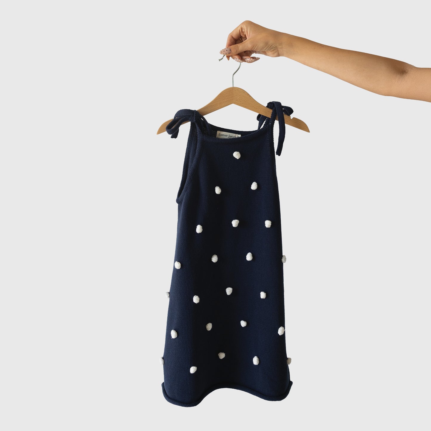 A hand holds the Knit Dress | Emery by forever french baby, a navy blue sleeveless 100% cotton dress with white pom-pom details and adjustable tie straps, displayed on a wooden hanger against a plain light background.