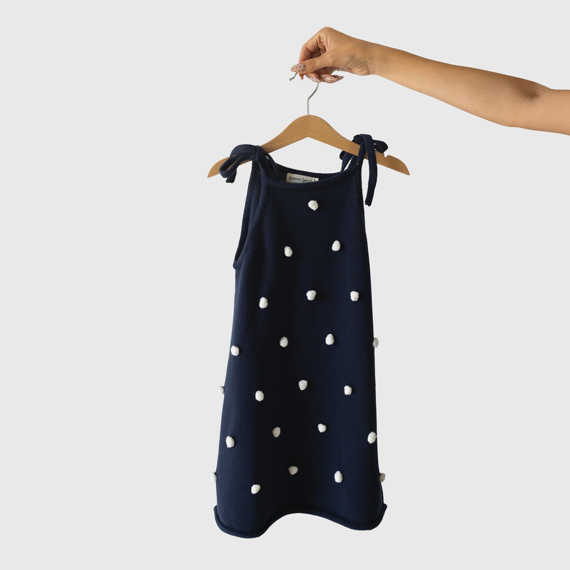 A hand holds the Knit Dress | Emery by forever french baby, a navy blue sleeveless 100% cotton dress with white pom-pom details and adjustable tie straps, displayed on a wooden hanger against a plain light background.
