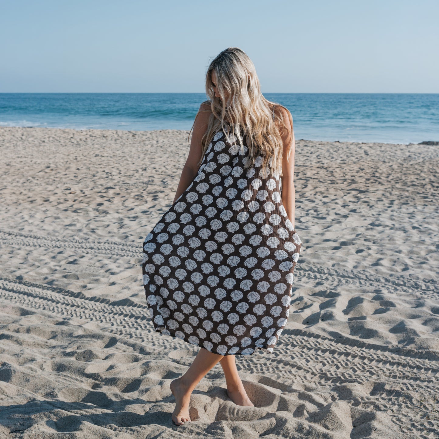 A woman with long blonde hair stands barefoot on a sandy beach, wearing the Women's Muslin Sleeveless Dress | Seashells by forever french baby. The ocean and blue sky complete this perfect scene for stylish coastal family outfits.