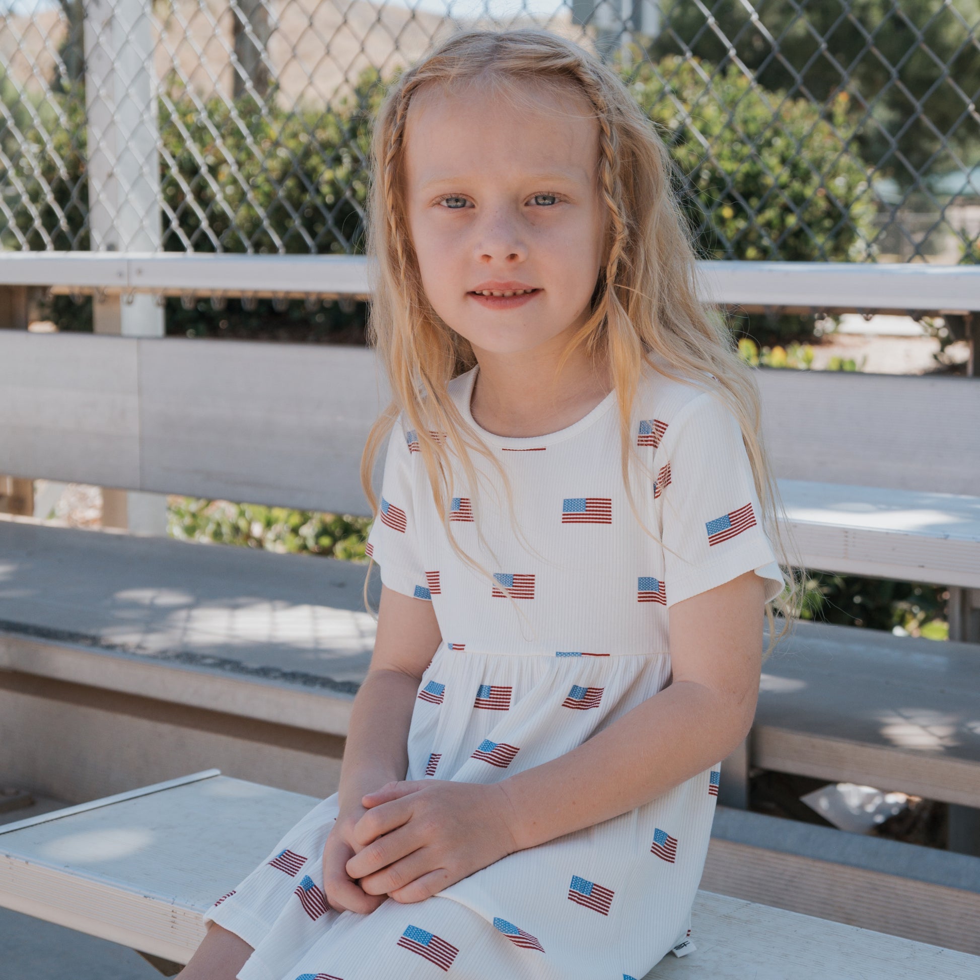 A young girl with long blonde hair sits on outdoor bleachers, wearing the Bamboo T-Shirt Dress | Americana Ribbed by forever french baby—a charming example of hypo-allergenic clothing. A chain-link fence and greenery are in the background.