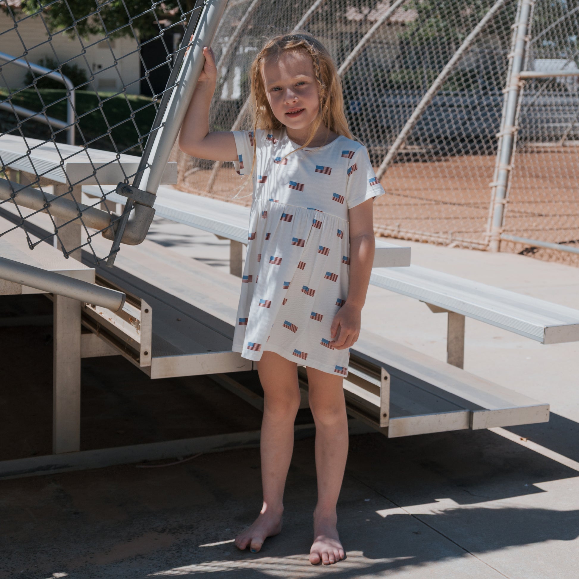 A young girl stands barefoot on bleachers, holding a fence and smiling slightly. She wears the forever french baby Bamboo T-Shirt Dress | Americana Ribbed outdoors on a sunny day.